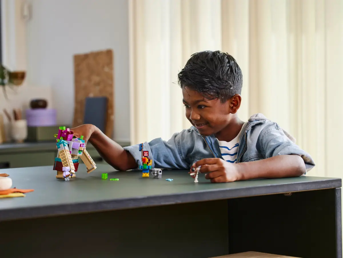 Uśmiechnięty chłopiec bawi się figurkami Lego na stole. Kolorowe konstrukcje Lego są przed nim. Tło obejmuje szafkę.