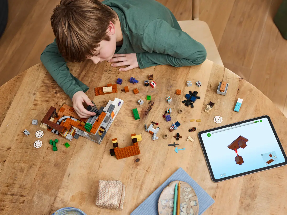Dziecko budujące zestaw Lego na drewnianym stole, z tabletem w pobliżu.