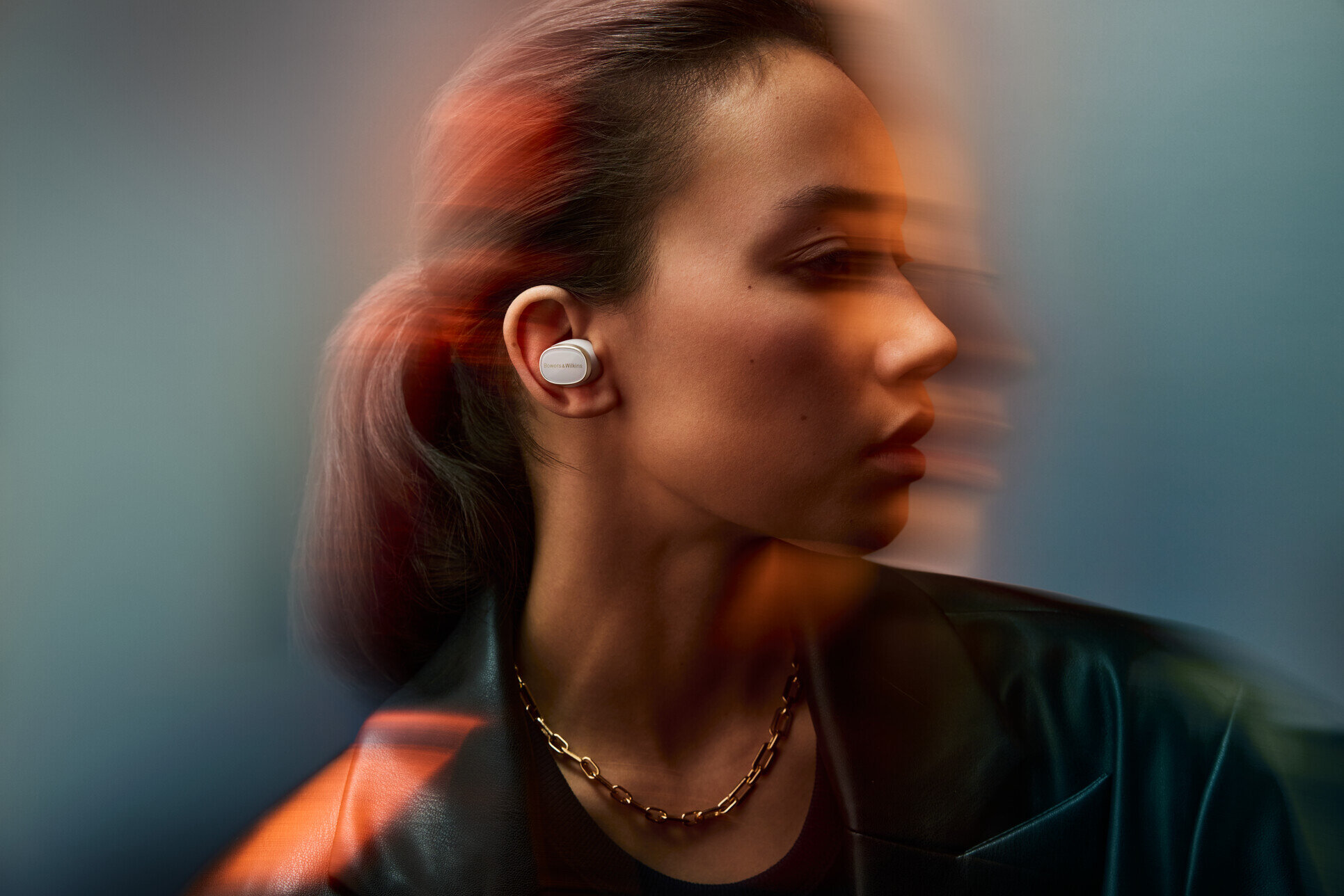 Kobieta ze słuchawkami. Ma na sobie czarną kurtkę i złoty naszyjnik. Tło z efektem rozmycia ruchu.