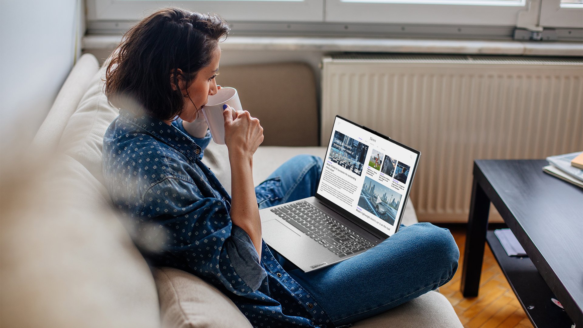 Frau auf Sofa mit Laptop, Becher und Fenster. Sie liest Nachrichtenartikel auf dem Laptop-Bildschirm.