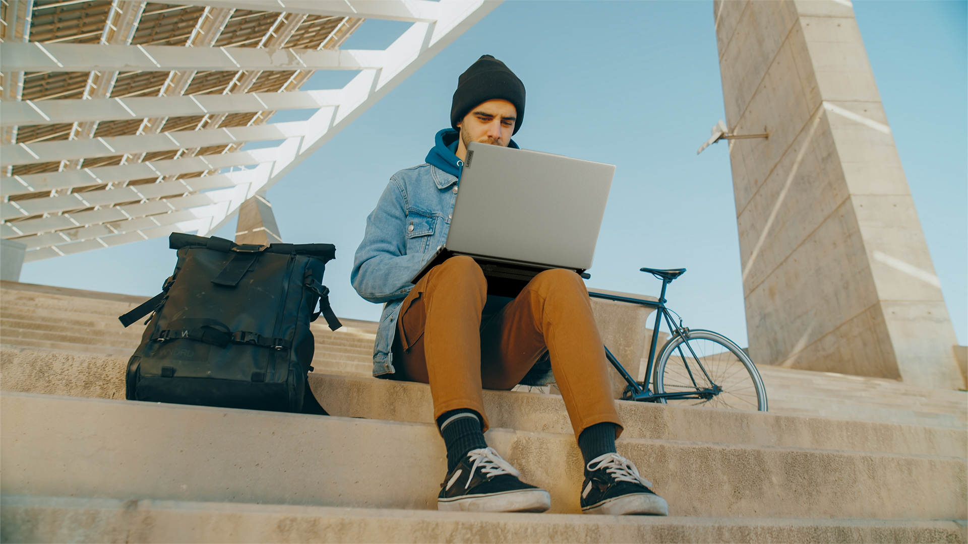 Mann sitzt auf Treppe und benutzt Laptop. Schwarzer Rucksack und Fahrrad in der Nähe.