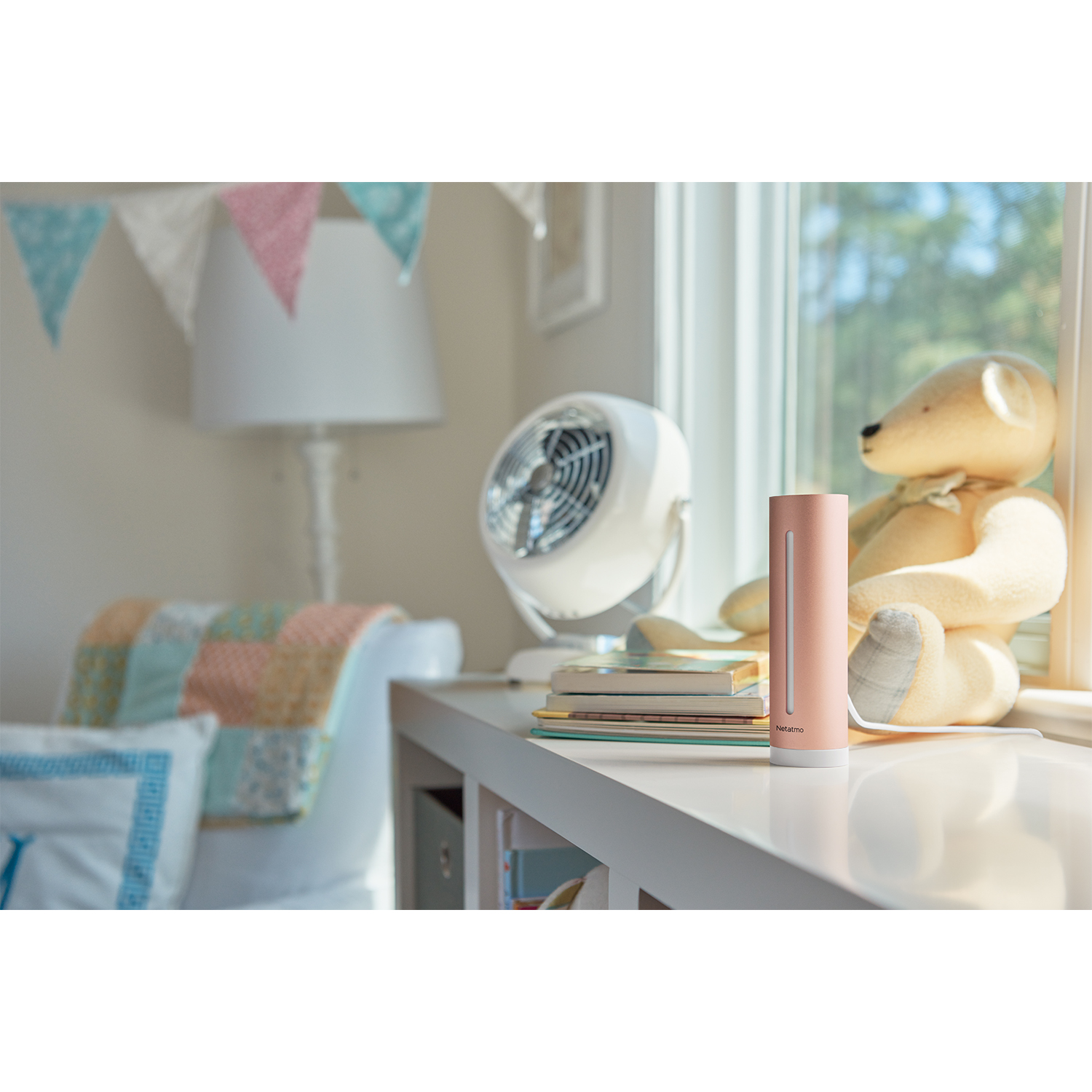 Un dispositivo rosa Netatmo smart home su un tavolo bianco vicino a una finestra, con un orsacchiotto e un ventilatore.