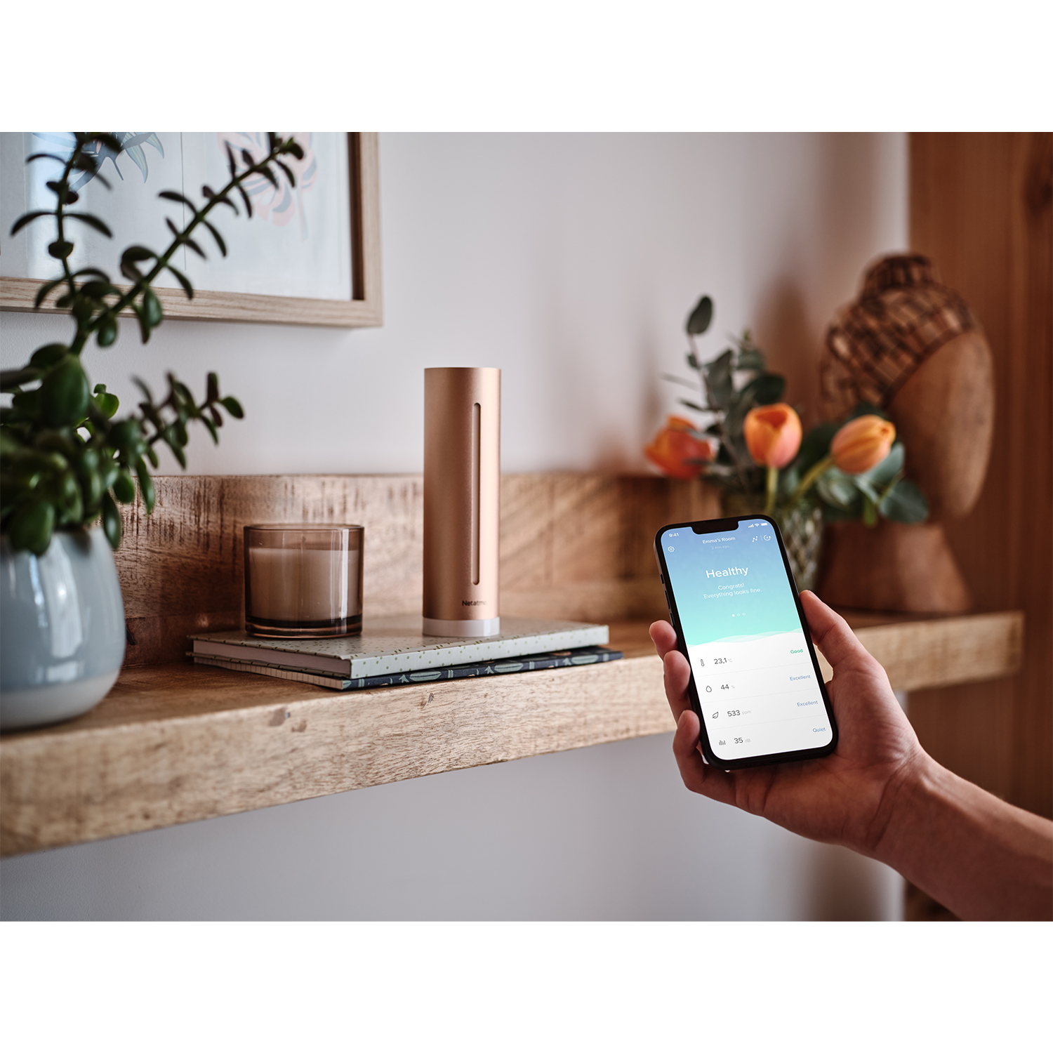 Una mano tiene un telefono su una mensola con un dispositivo intelligente, una candela e fiori.