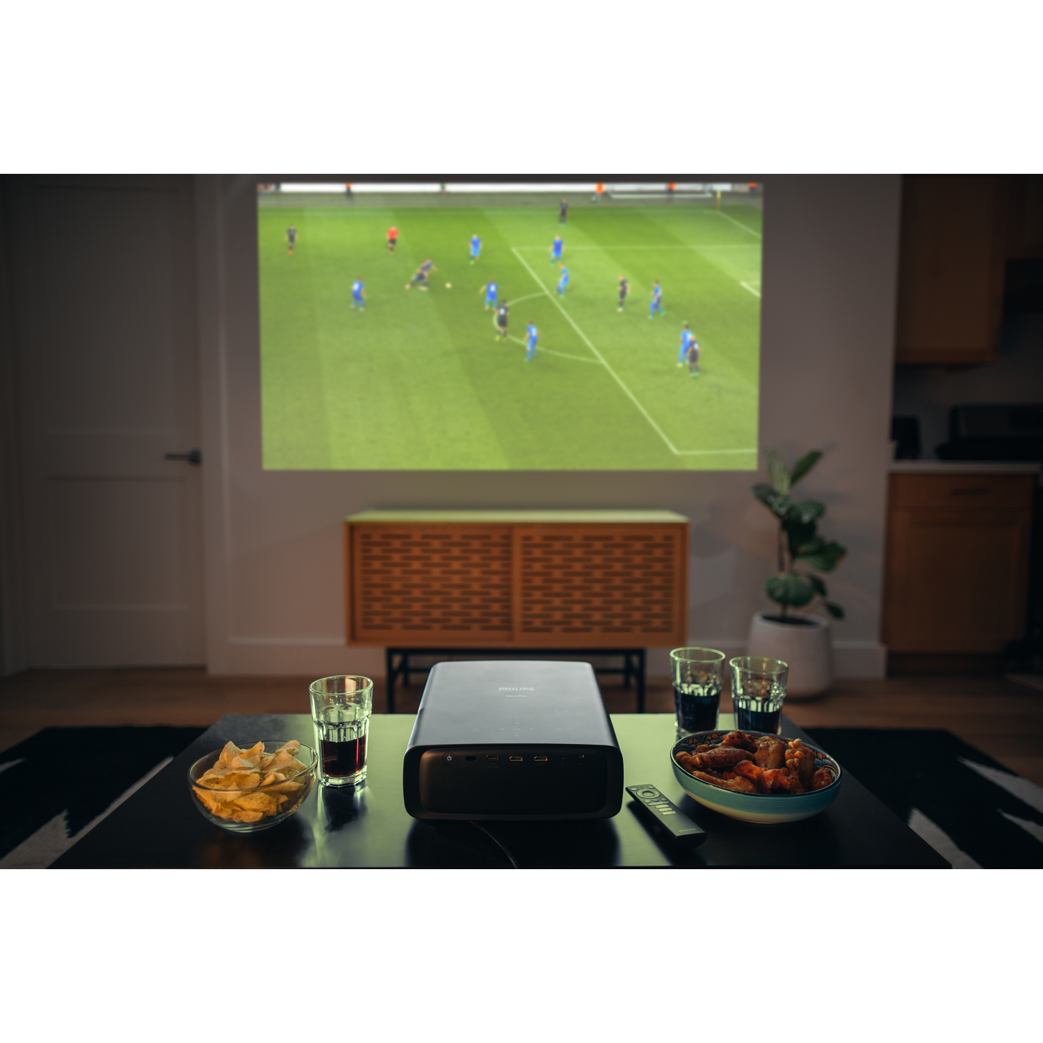 A projector displays a soccer game. Snacks and drinks sit on a table in front of the screen.