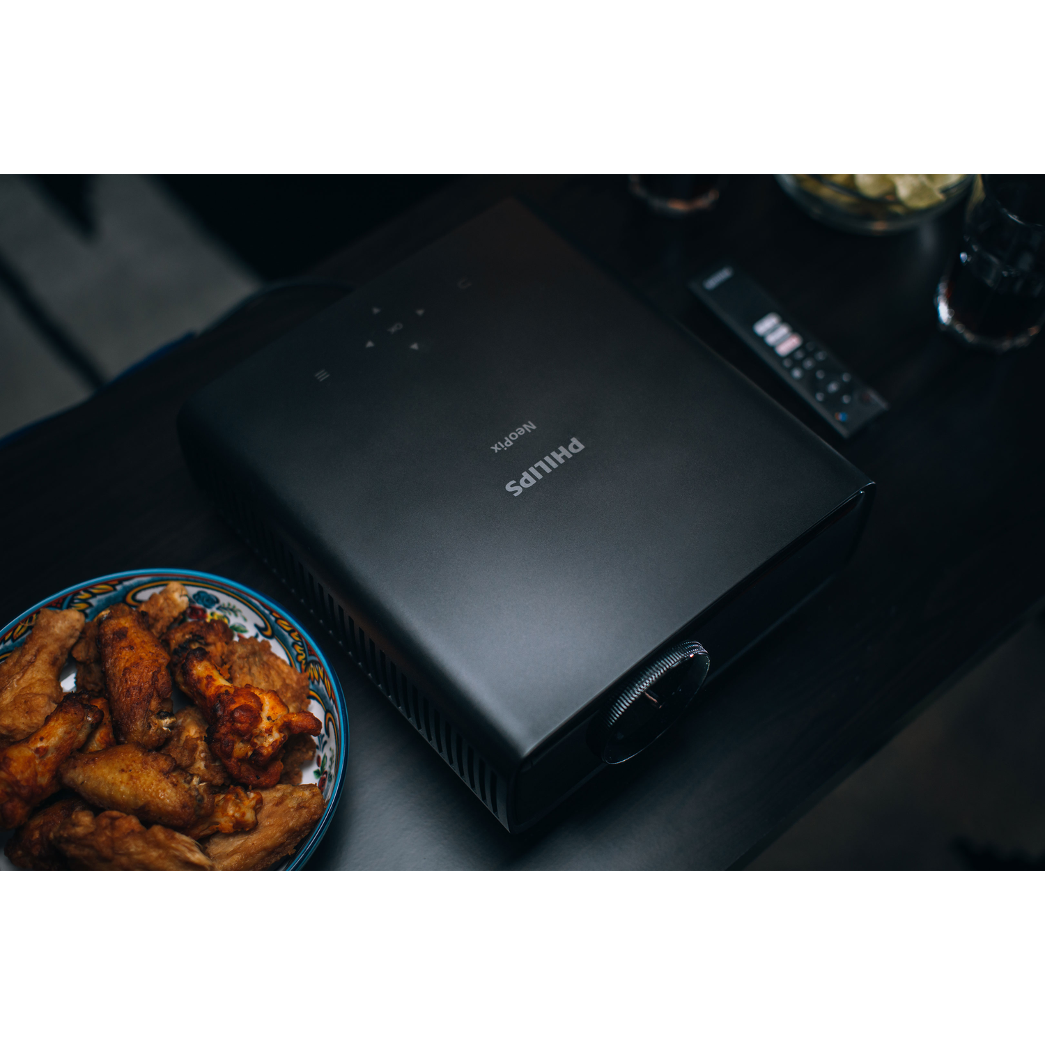 A black projector with a remote and a bowl of food on a table.