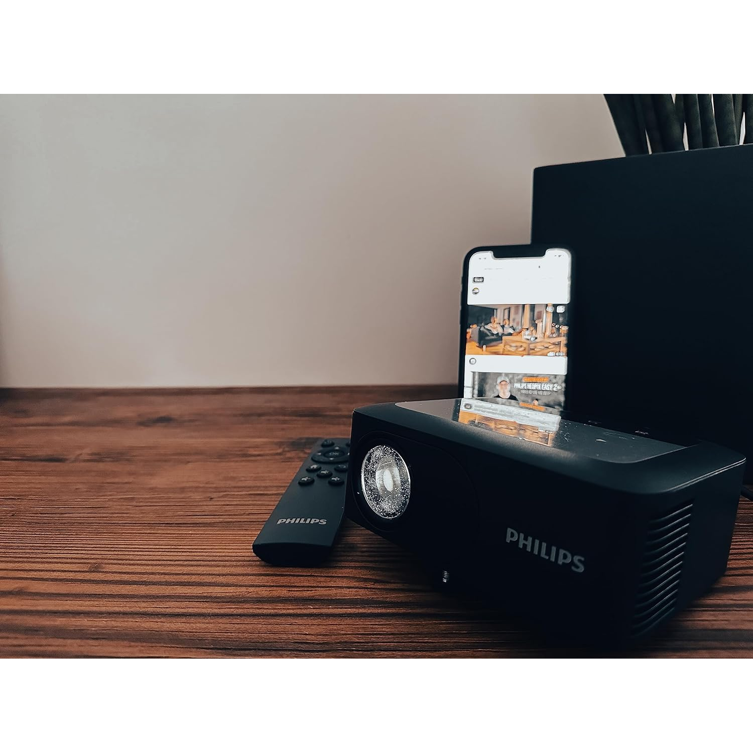 Black projector and remote control on a wooden table, with a phone and a black box in the background.