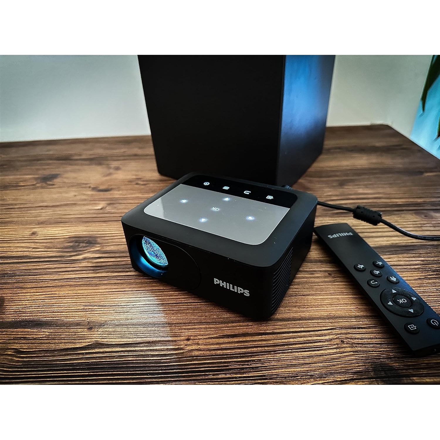 Black Philips projector and remote on a wooden table with a black speaker in background.