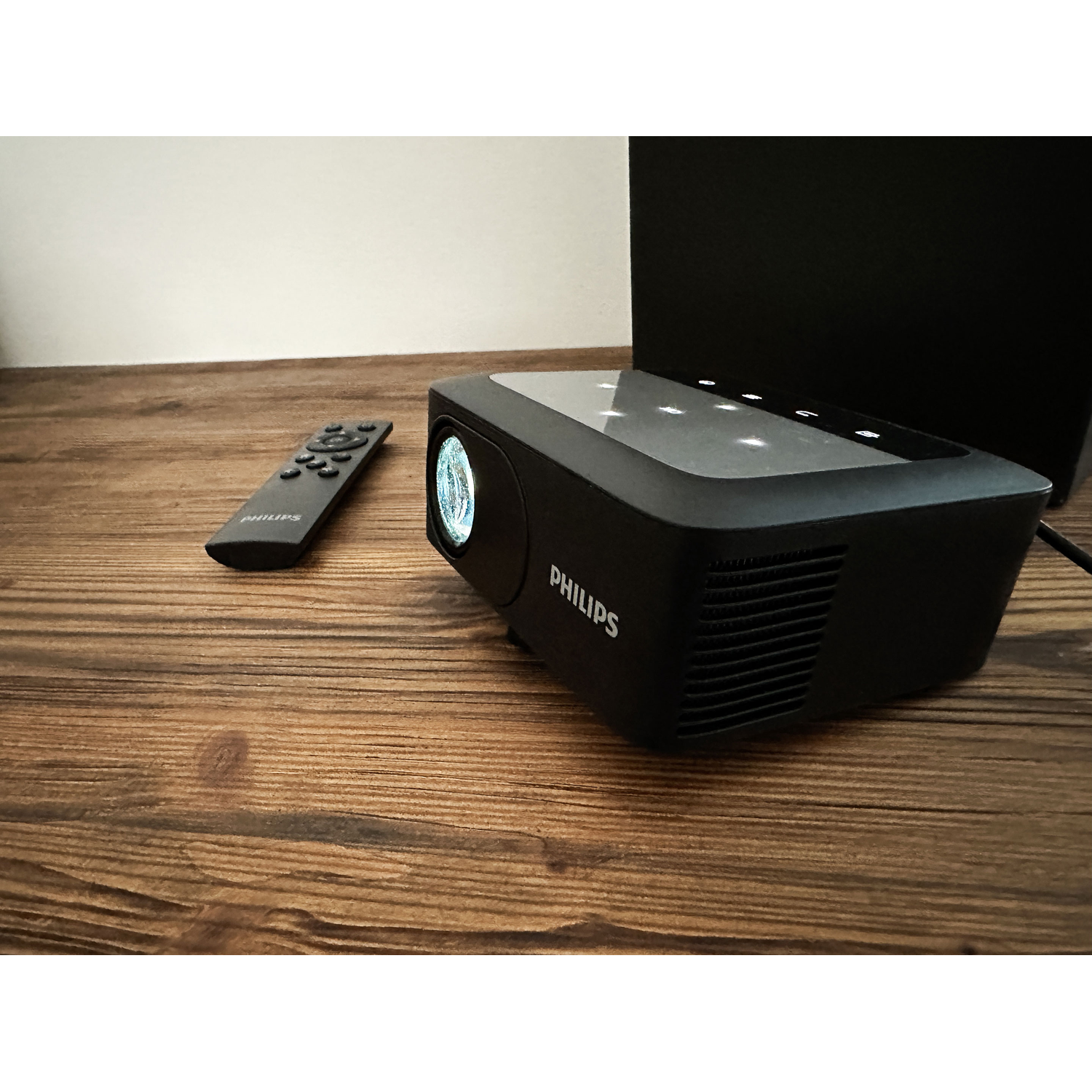 Black projector with remote on a wooden table. The word PHILIPS is visible.