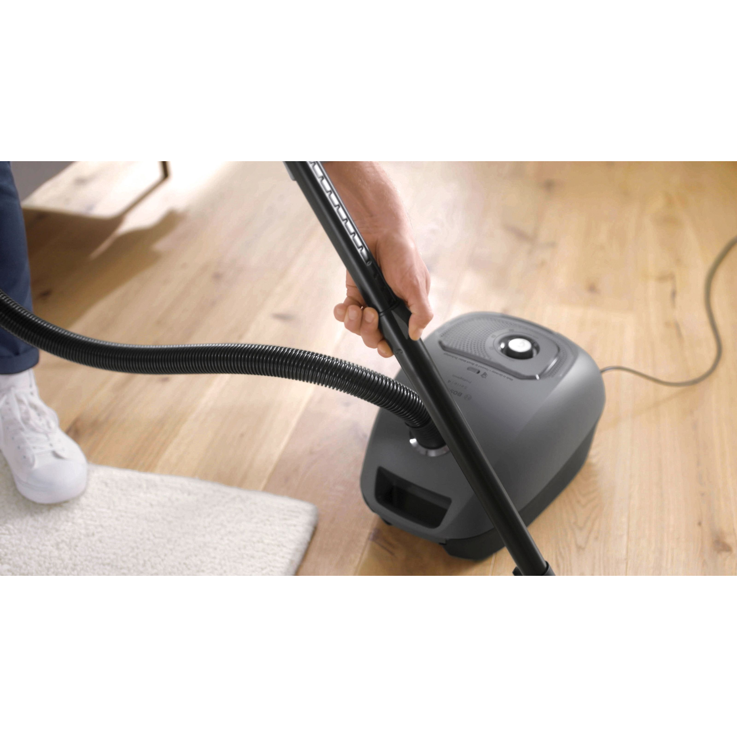 Person cleaning a wooden floor with a gray steam cleaner, white rug in the corner.
