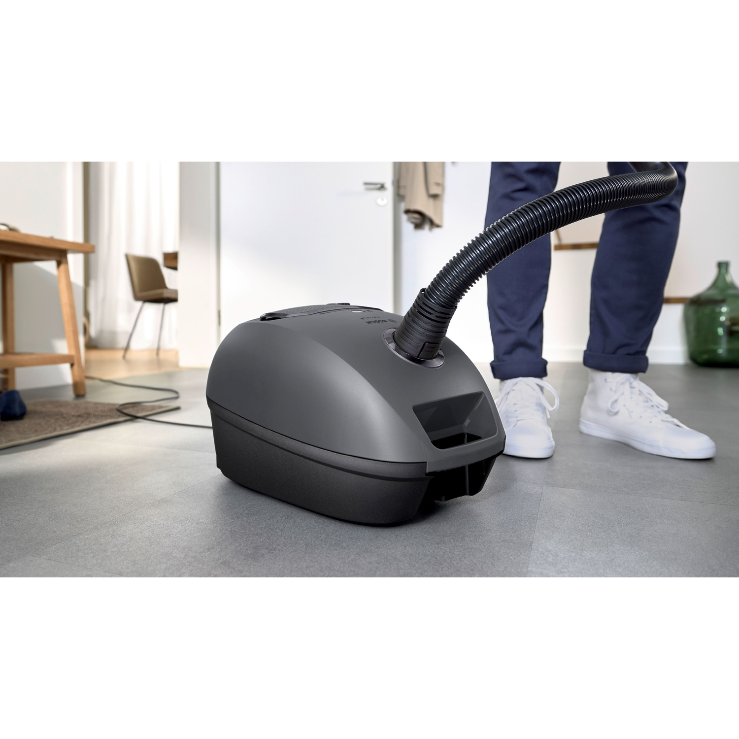 A person vacuuming a gray vacuum cleaner. The person wears jeans and white sneakers.