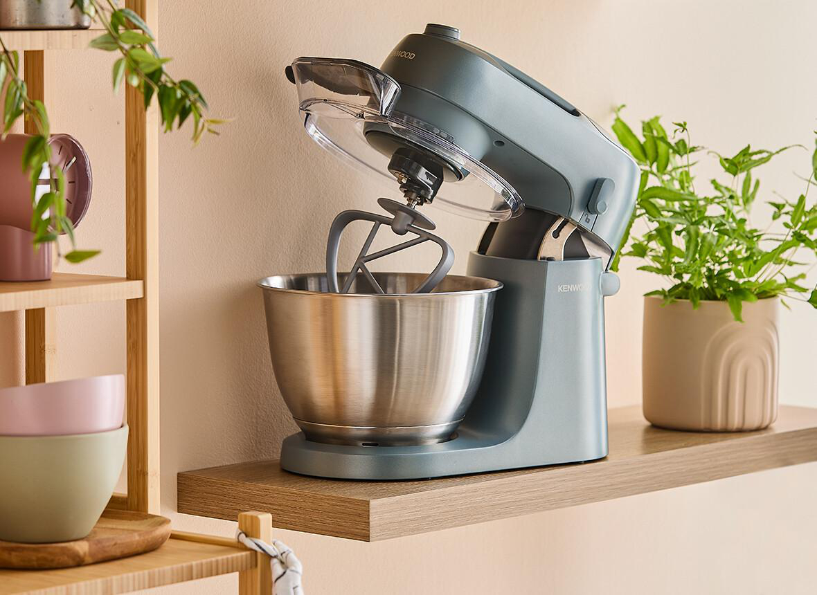 Ein grauer Küchenmixer auf einem Holzregal mit Pflanzen und Schalen.