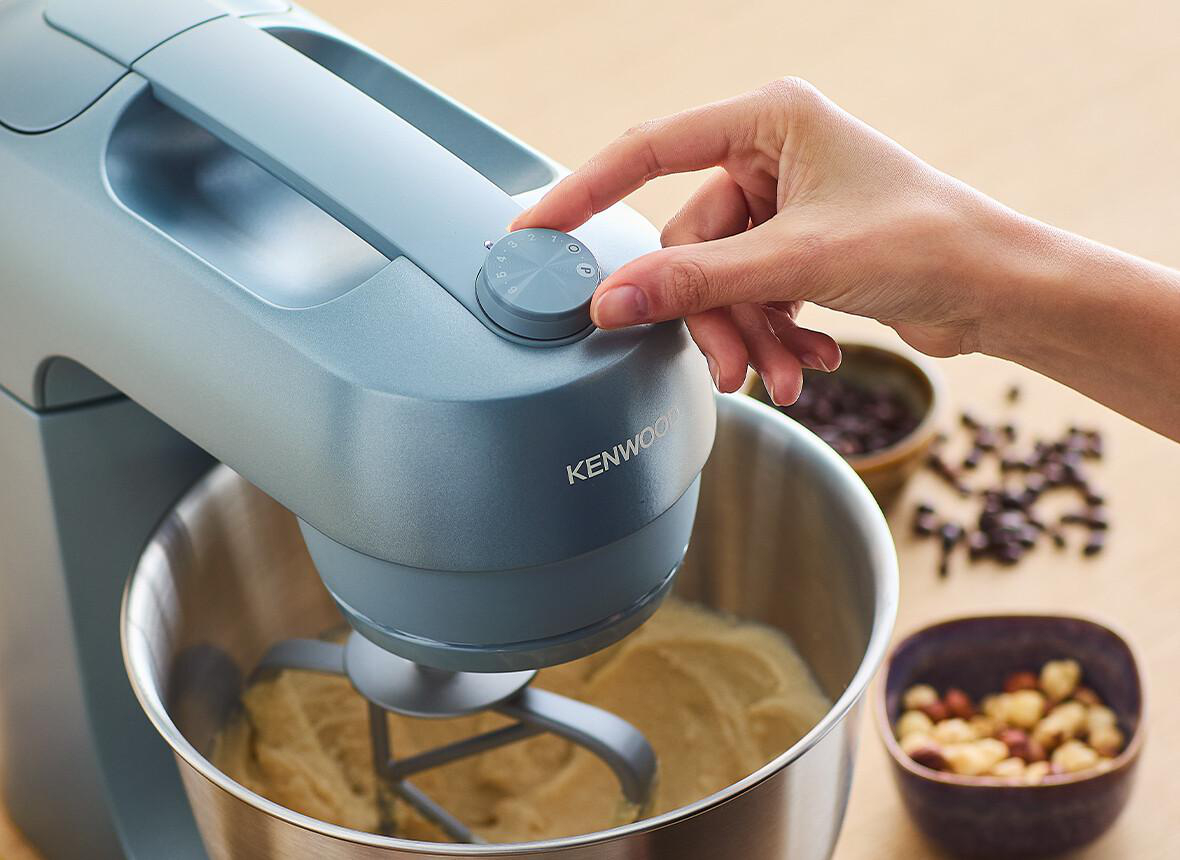 Hand dreht einen Mixer-Drehregler an einem Kenwood-Mixer, Zutaten im Hintergrund.