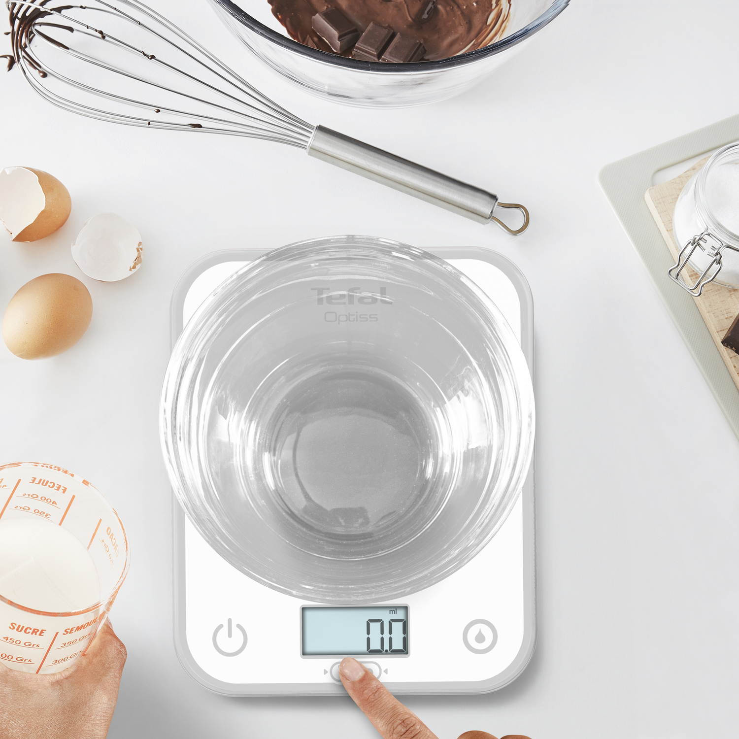 Une balance de cuisine avec un bol en verre et une main appuyant sur un bouton. Ingrédients et ustensiles de cuisine.