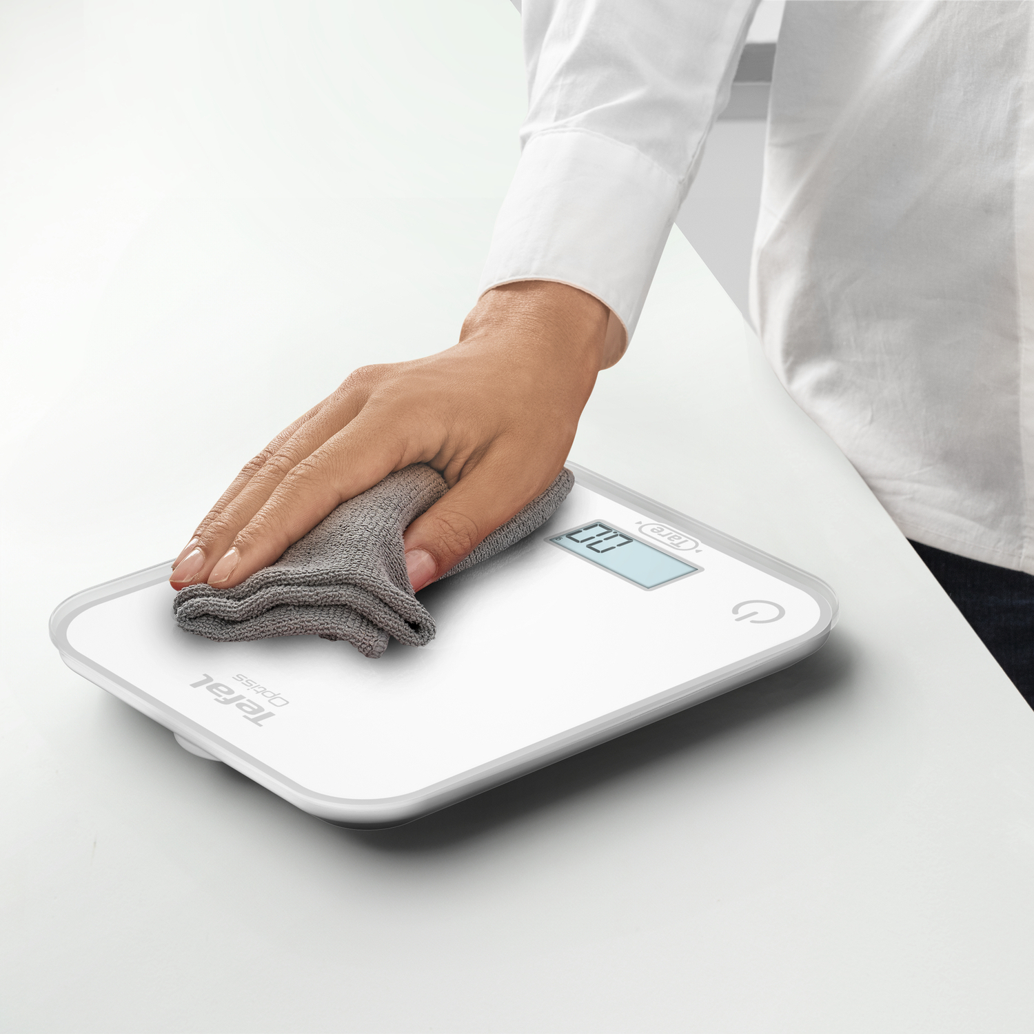 Une personne essuie une balance numérique blanche avec un chiffon gris sur une surface blanche.
