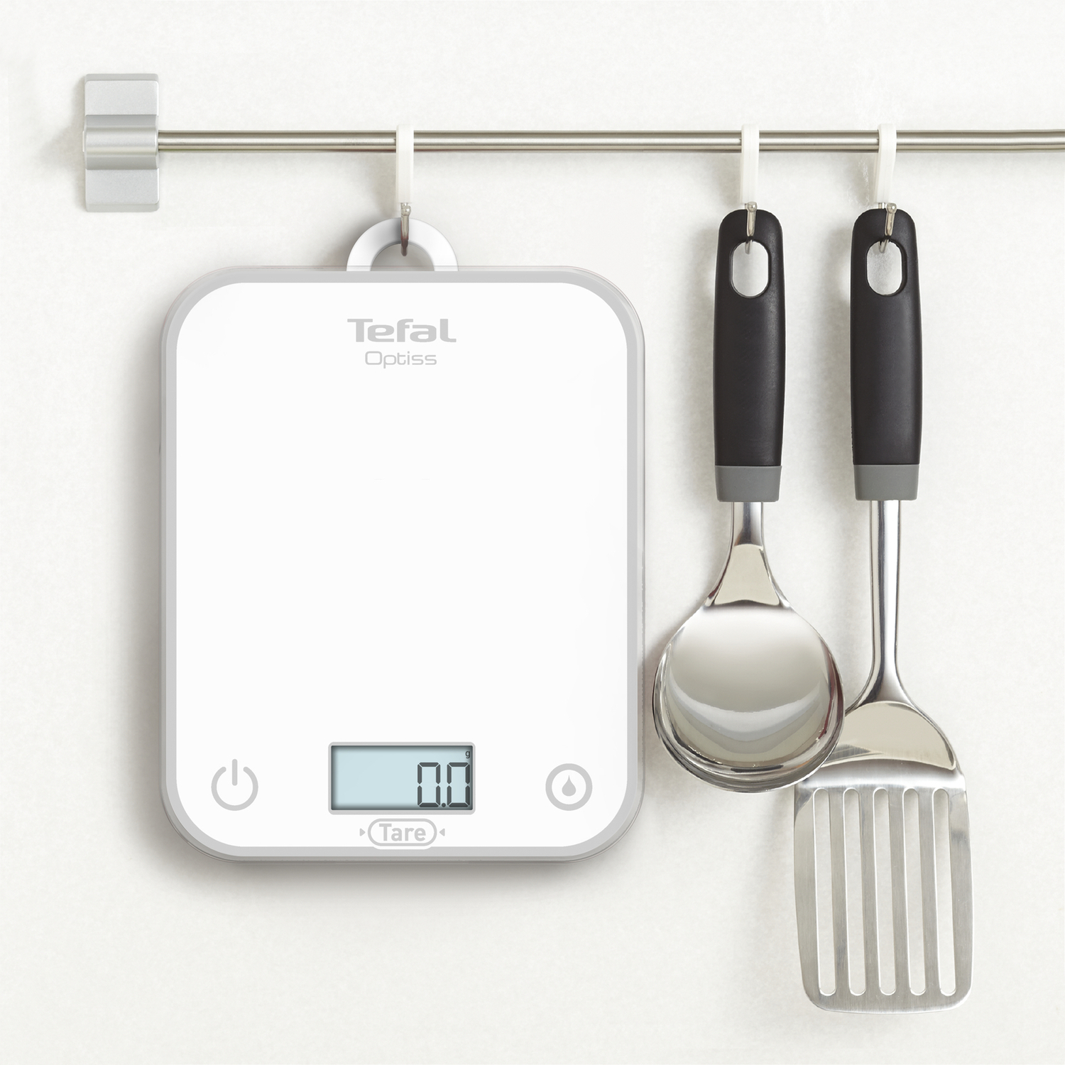 Balance de cuisine blanche, cuillères doseuses et spatule suspendues à une barre métallique sur mur blanc.