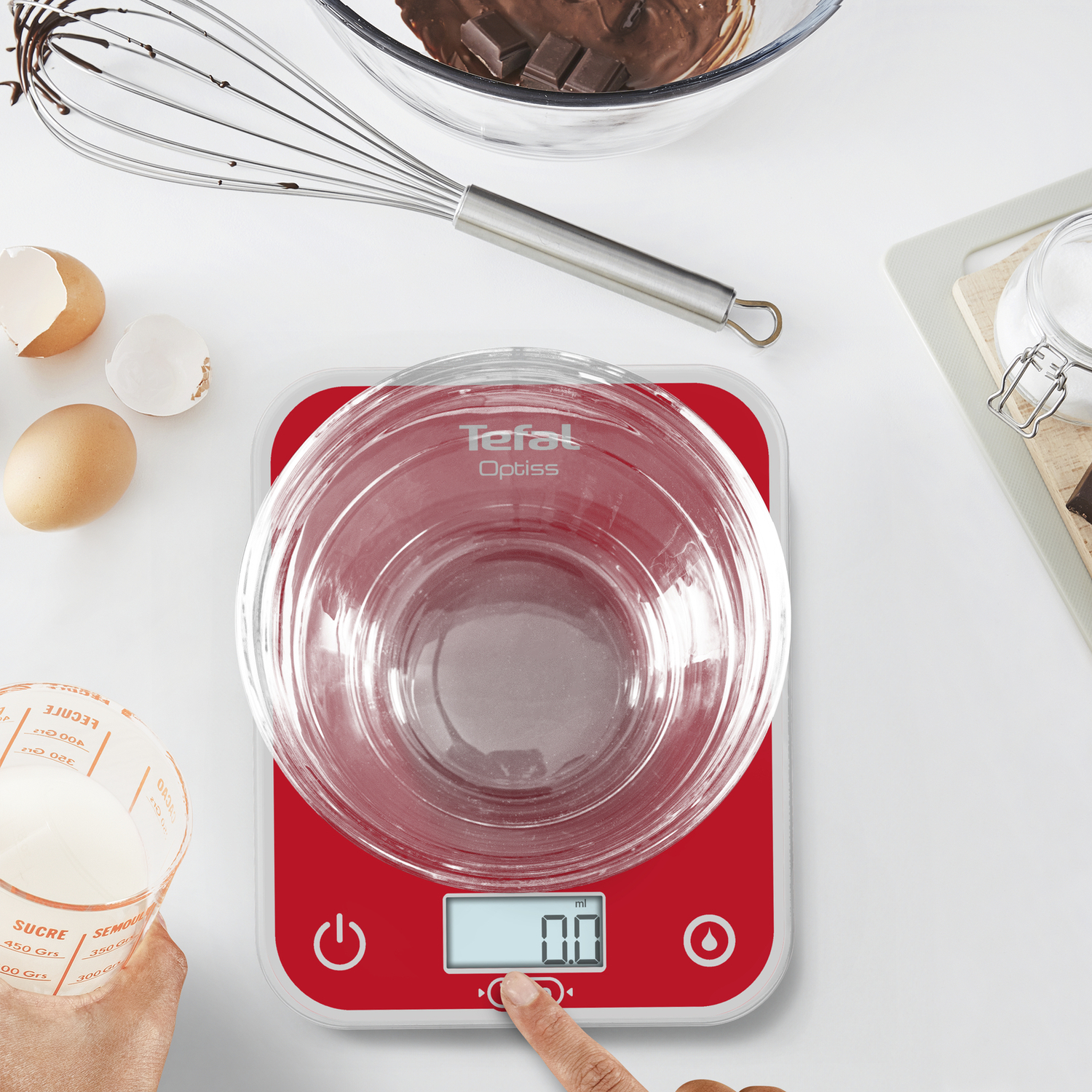 Balance de cuisine rouge et blanche avec un bol en verre et des ingrédients. Œufs, lait et chocolat visibles.