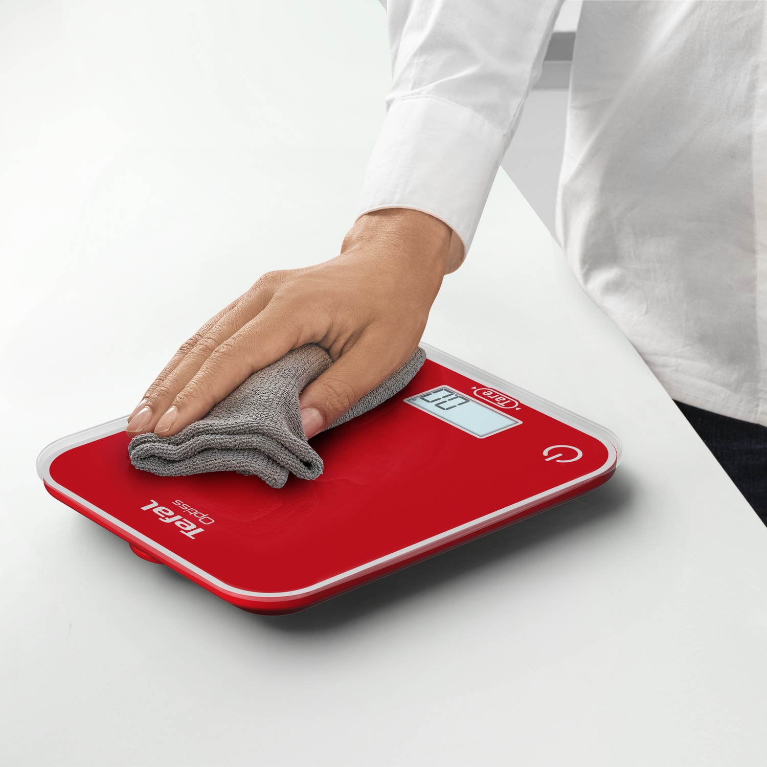 Main essuyant une balance numérique Tefal rouge avec un chiffon gris. La balance affiche '0.0'.
