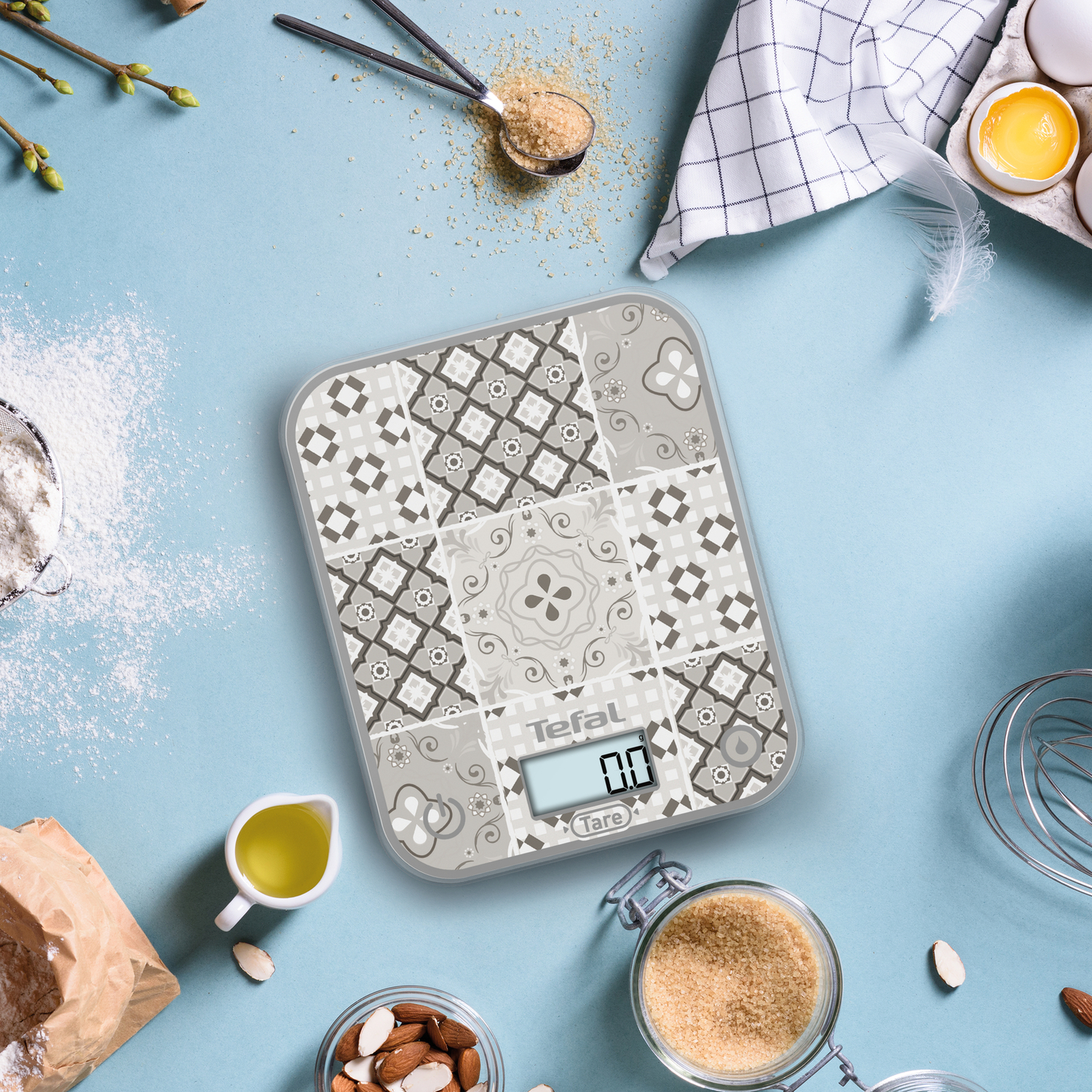 Balance de cuisine avec surface à motifs, entourée d'ingrédients comme de la farine, de l'huile et des amandes.