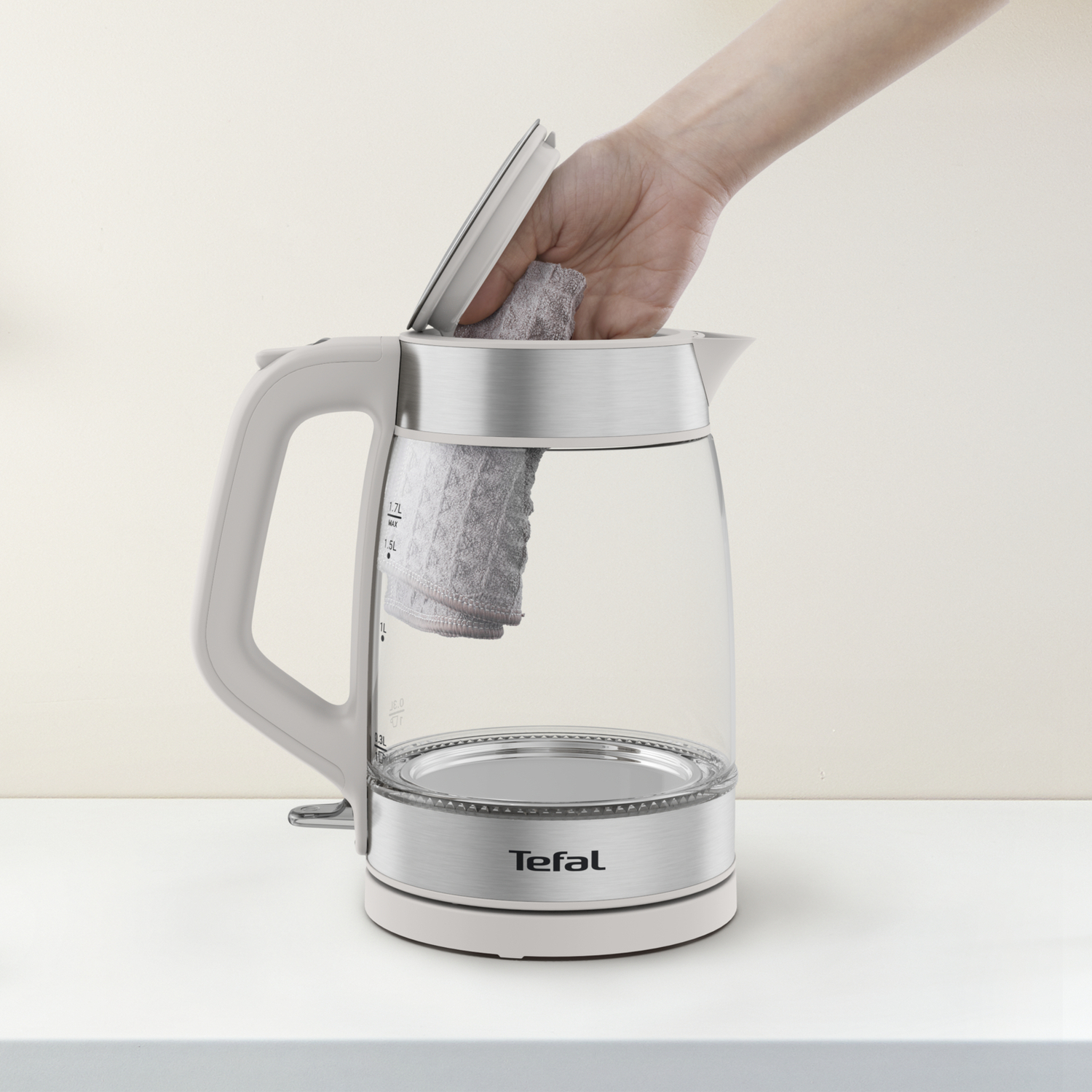A hand placing a cloth in a clear glass electric kettle. The kettle has a white base and handle.