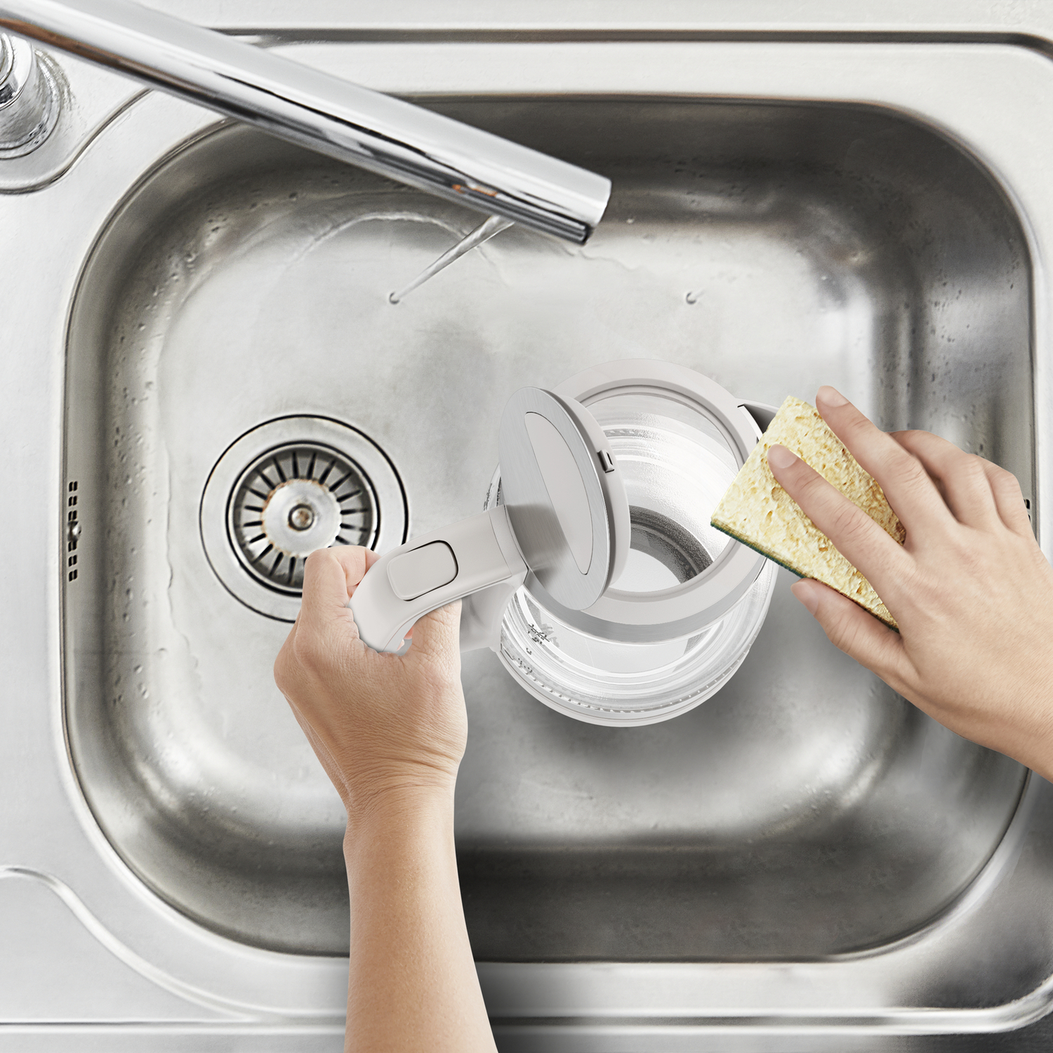 Hands washing a glass kettle in a stainless steel sink, with a sponge.