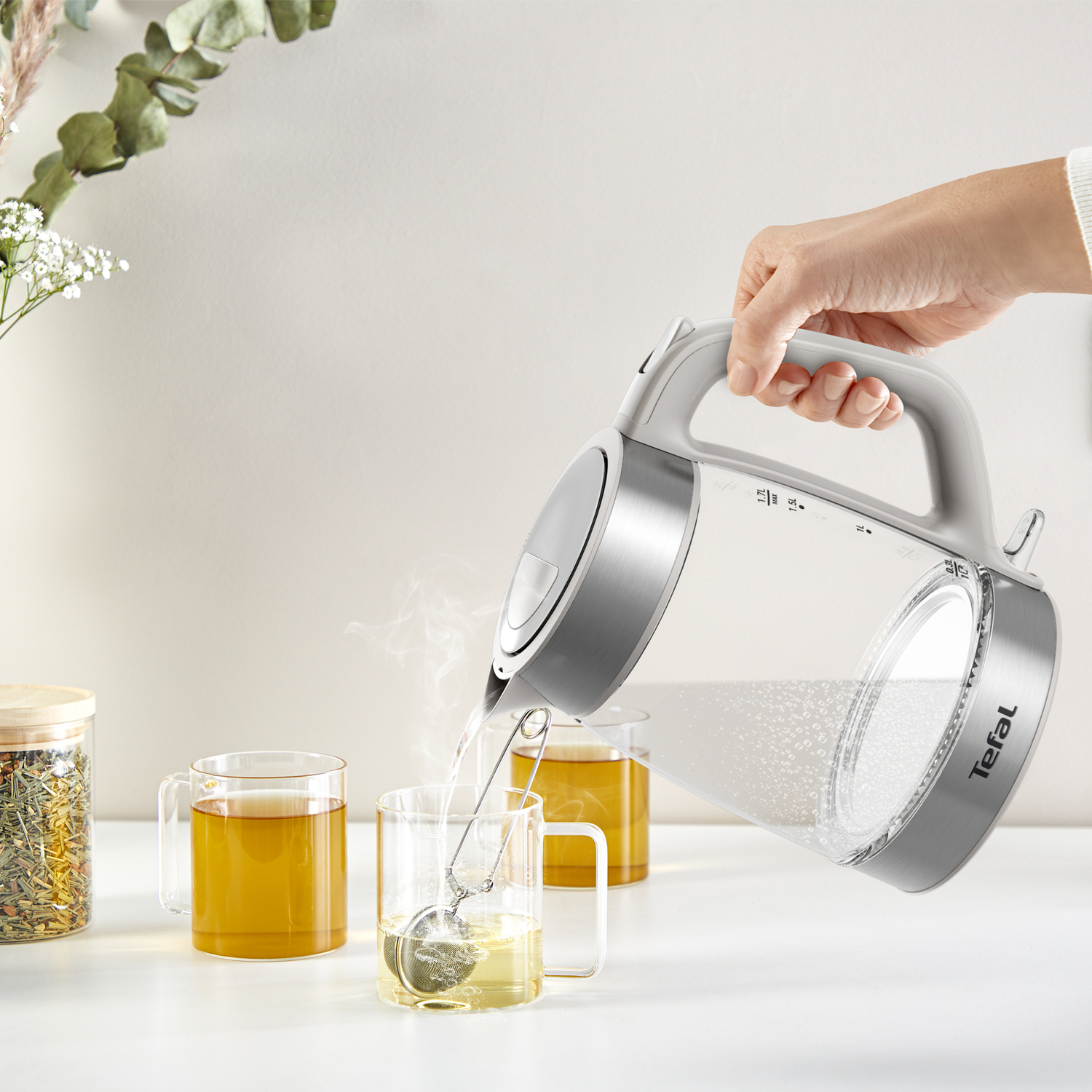 A hand pouring hot water from a silver kettle into a glass mug with tea, steam rising.