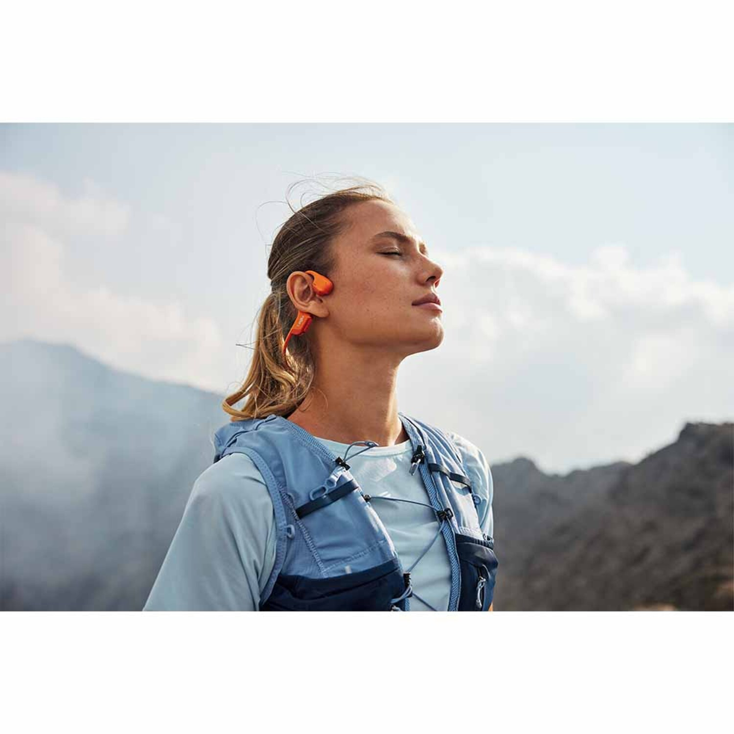 Vrouw met oranje sporthoofdtelefoon, blauw vest, buitenachtergrond.