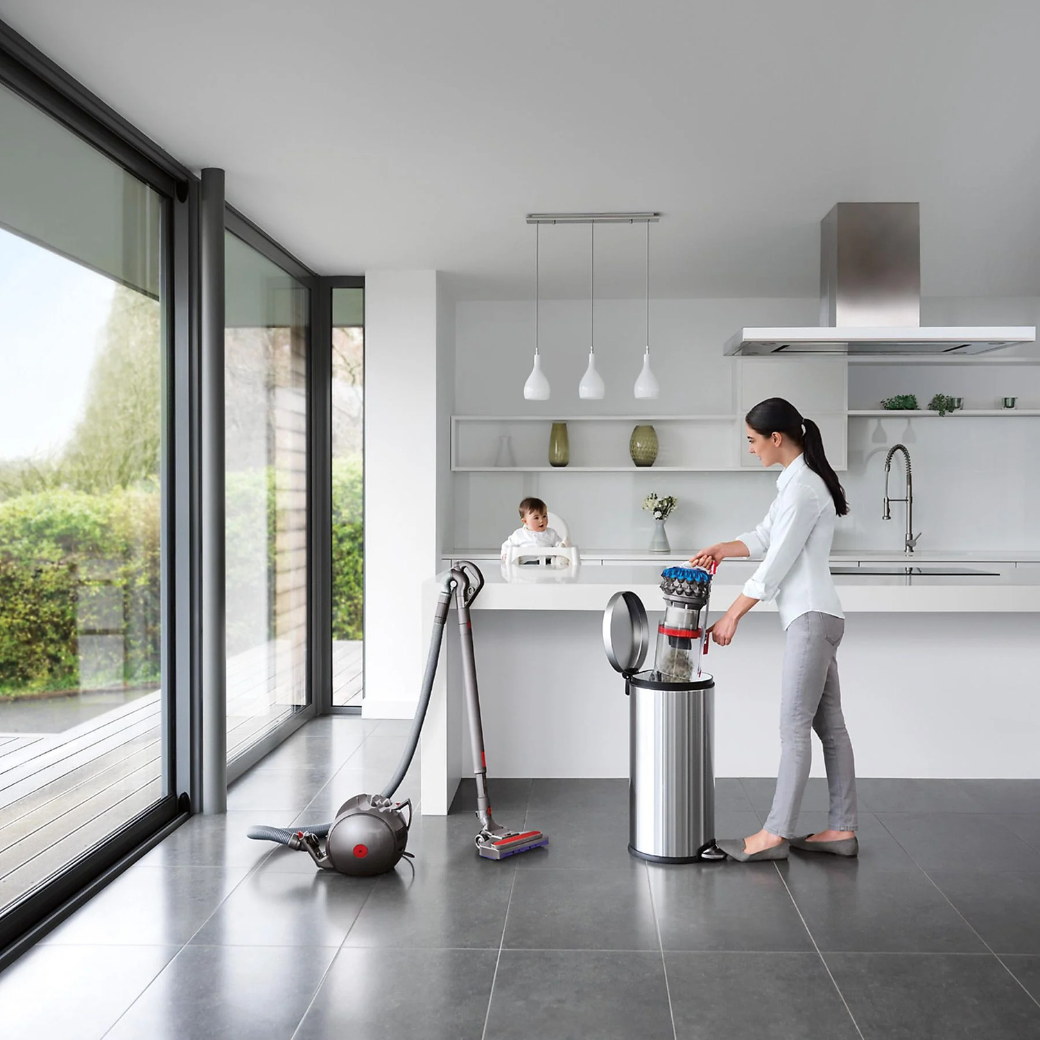 A woman in a white kitchen is throwing trash into a silver trash can.