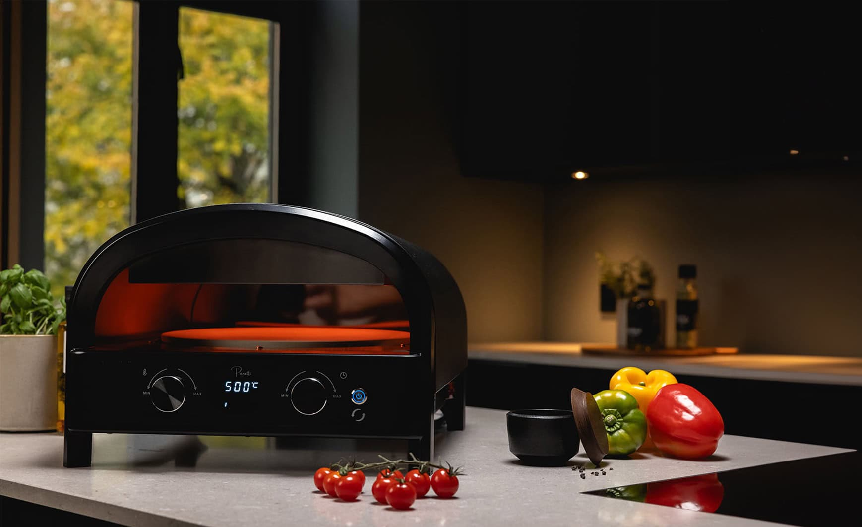 Fekete pizzasütő piros belsővel és pizzakővel, zöldségekkel körülvéve az asztalon.