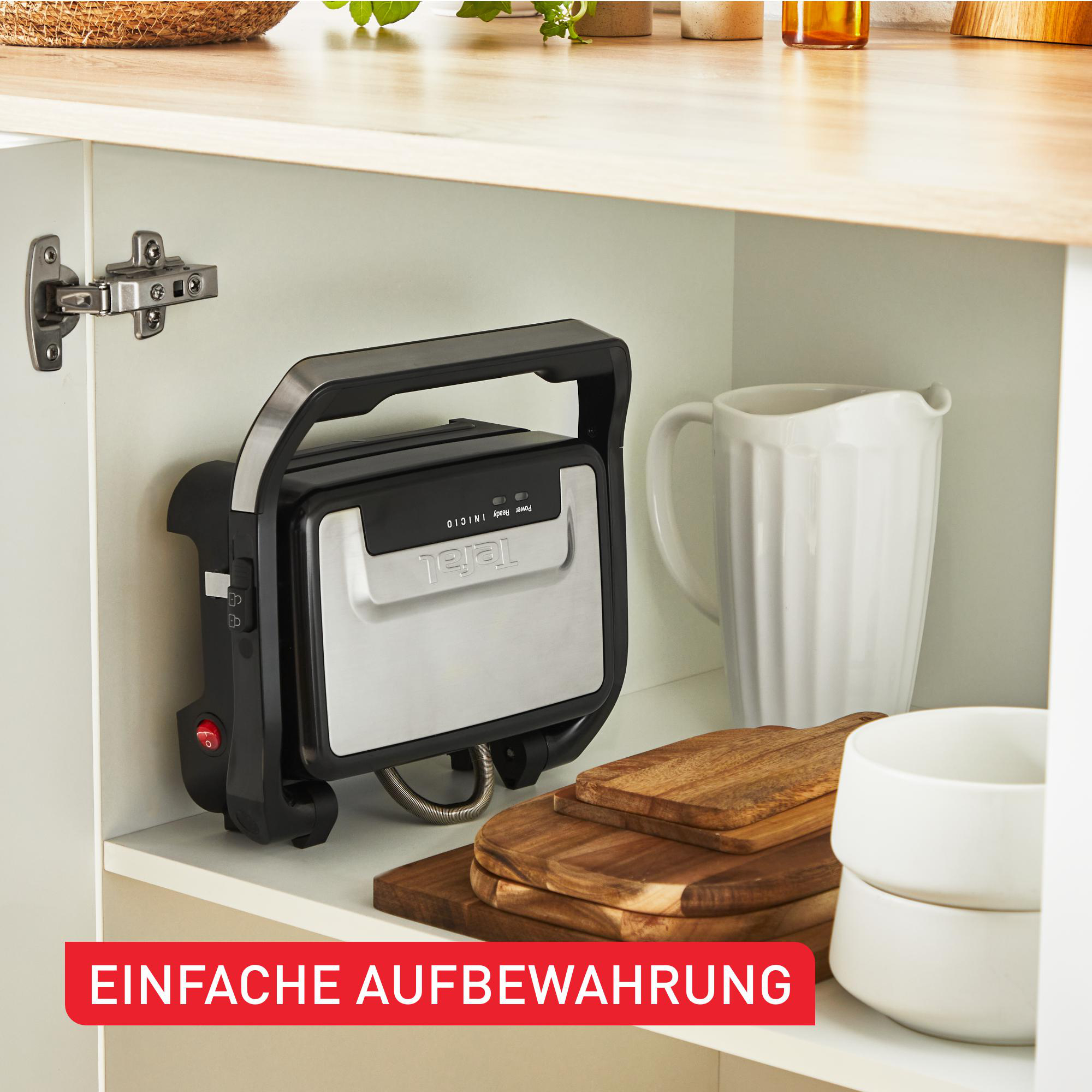 Ein schwarzer und silberner Tefal-Grill in einem weißen Schrank, mit Holzschneidebrettern und Tassen.