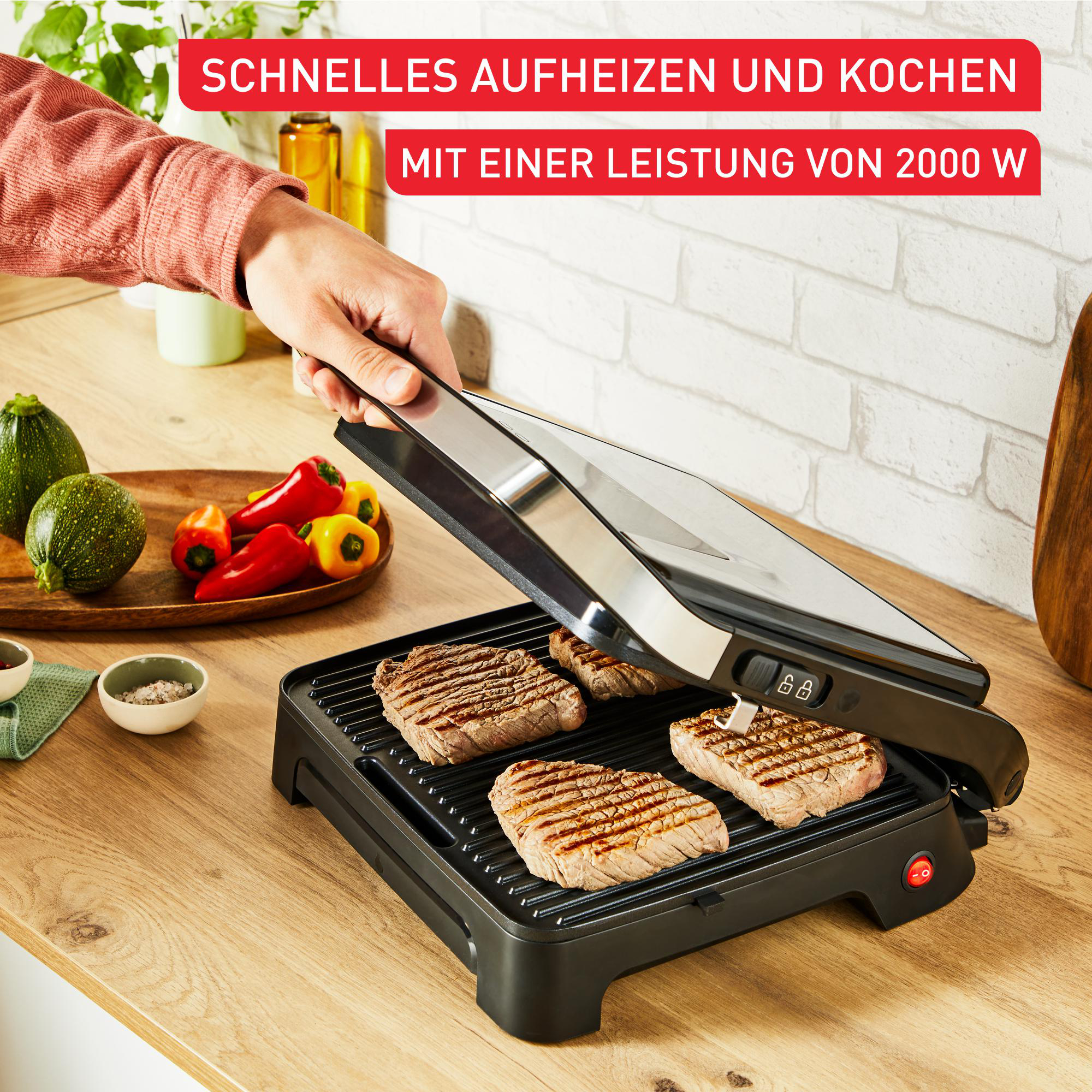Eine Person grillt Steaks auf einem schwarzen Grill mit Griff. Gemüse ist in der Nähe.