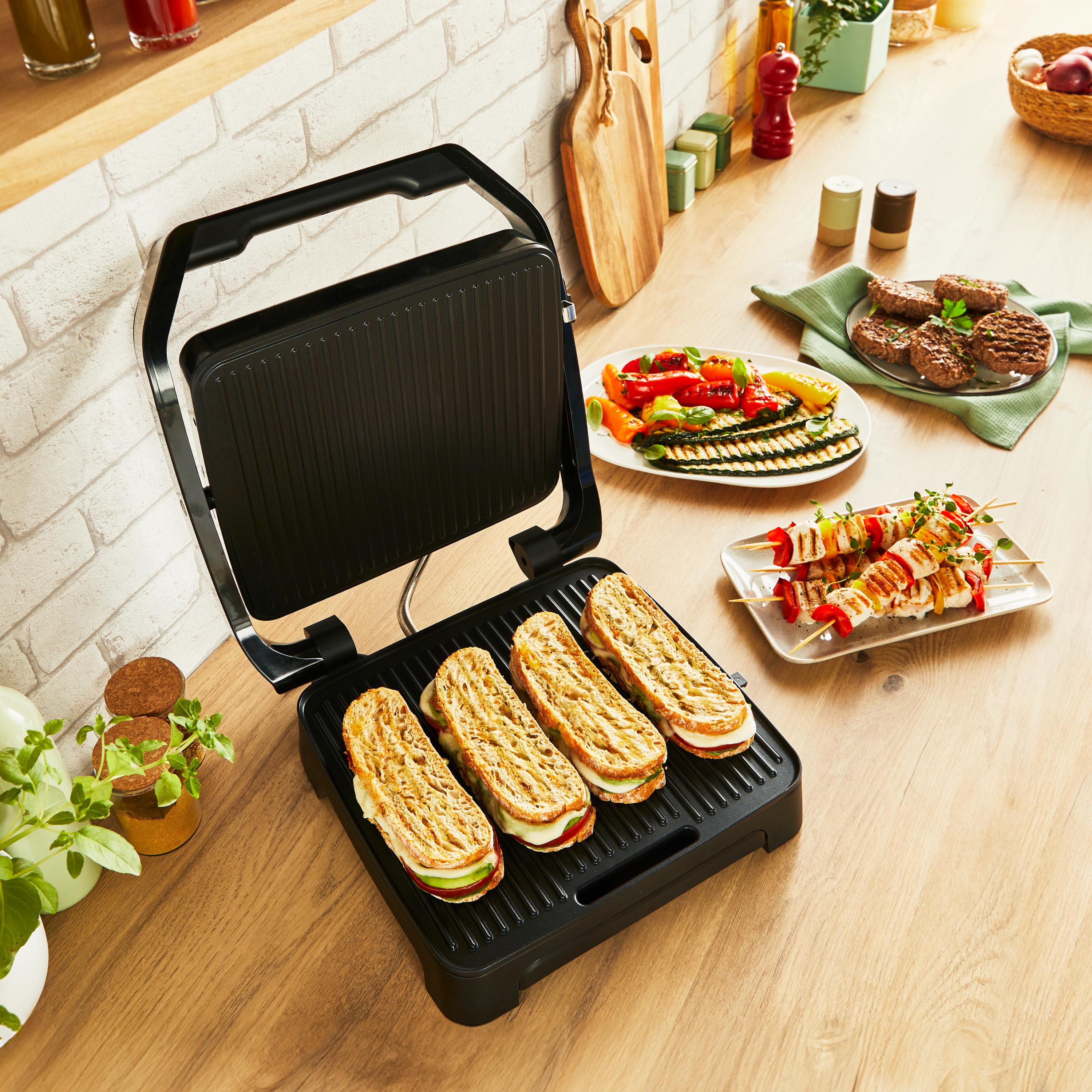 Grill mit Sandwiches, Gemüse und Spießen. Holztisch mit Essen. Hintergrund Backsteinmauer.