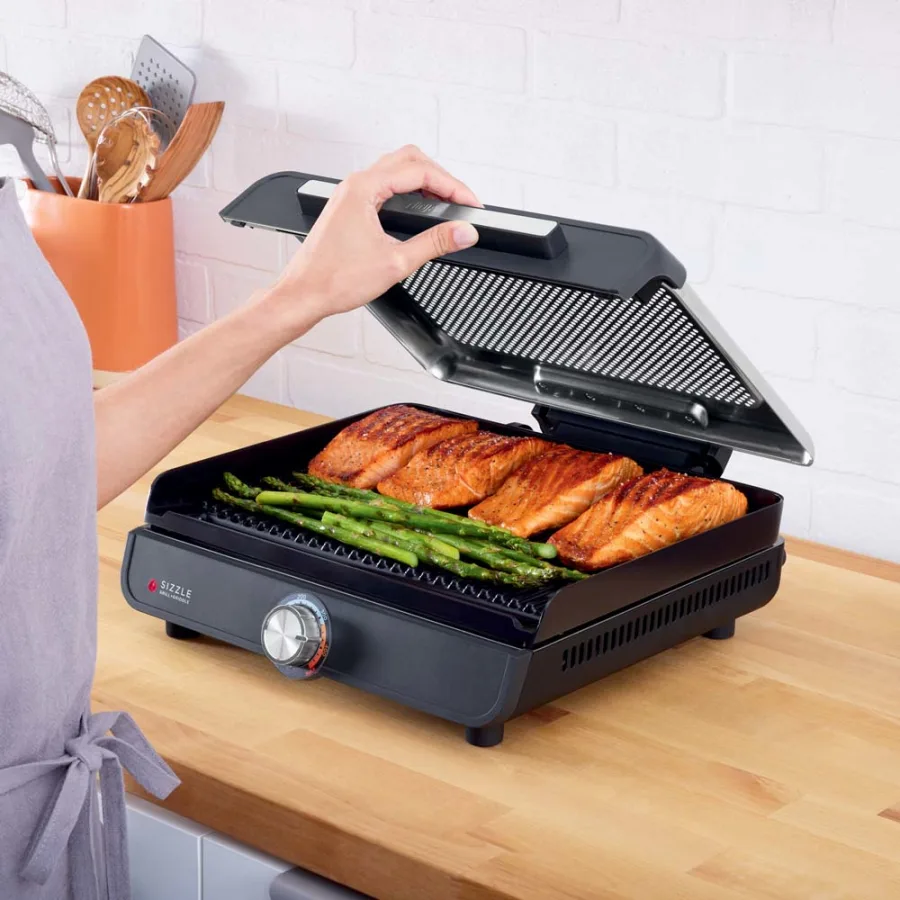 Egy személy lazacot és spárgát grillez beltéri grillen. A grill egy fa munkalapon van.