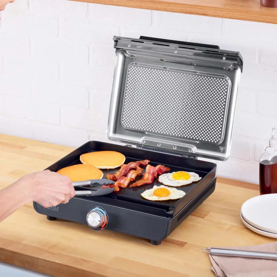 Személy csipesszel fordítja az ételt a grillen, szalonna, tojás és zsemle látható.