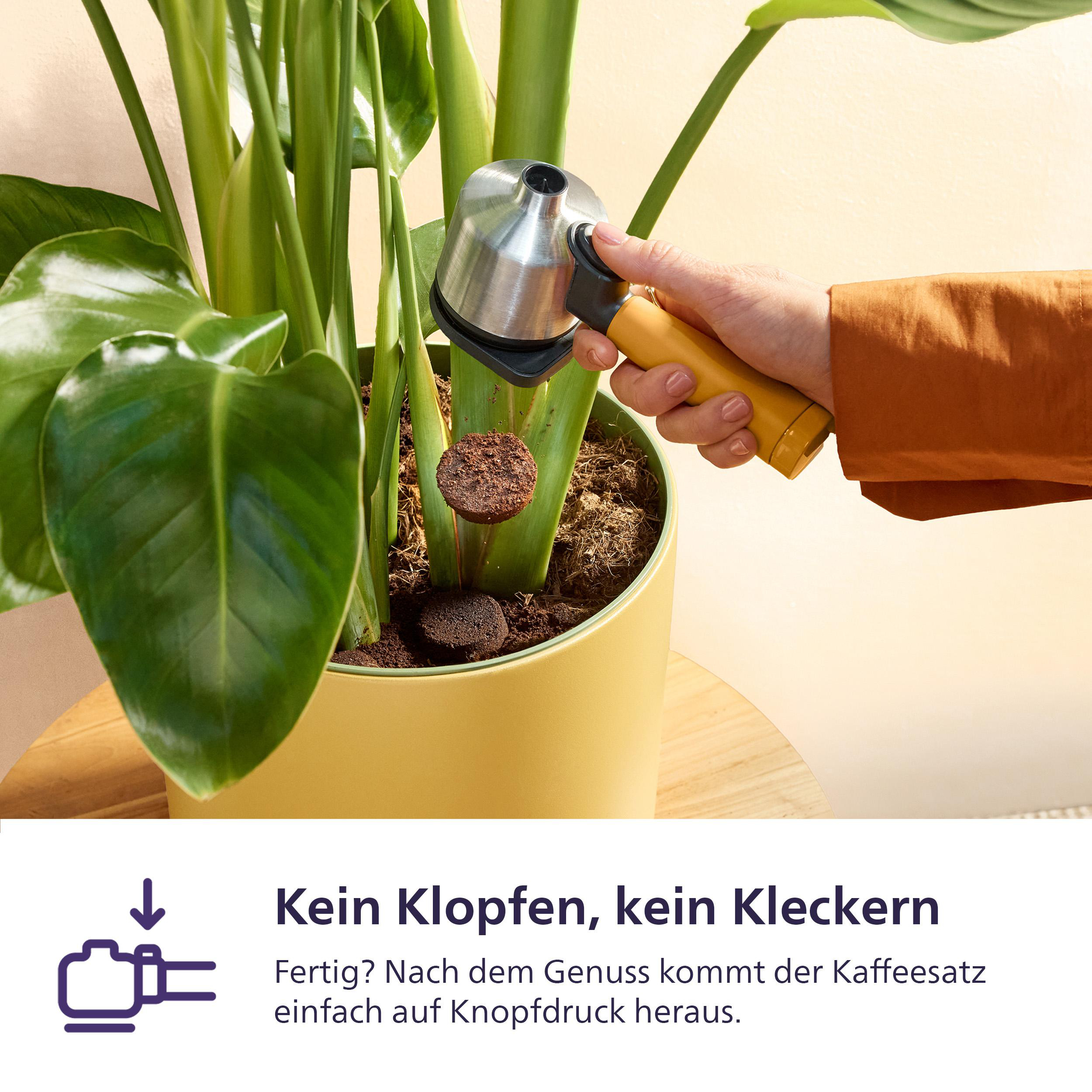 Eine Hand benutzt ein Werkzeug, um Kaffeesatz aus einem Blumentopf zu drücken. Grüne Blätter und ein gelber Topf sind sichtbar.