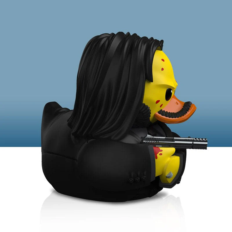 Egy John Wick-nek öltözött gumikacsa, fegyverrel a kezében. Sárga kacsa fekete öltönyben és hosszú hajjal.