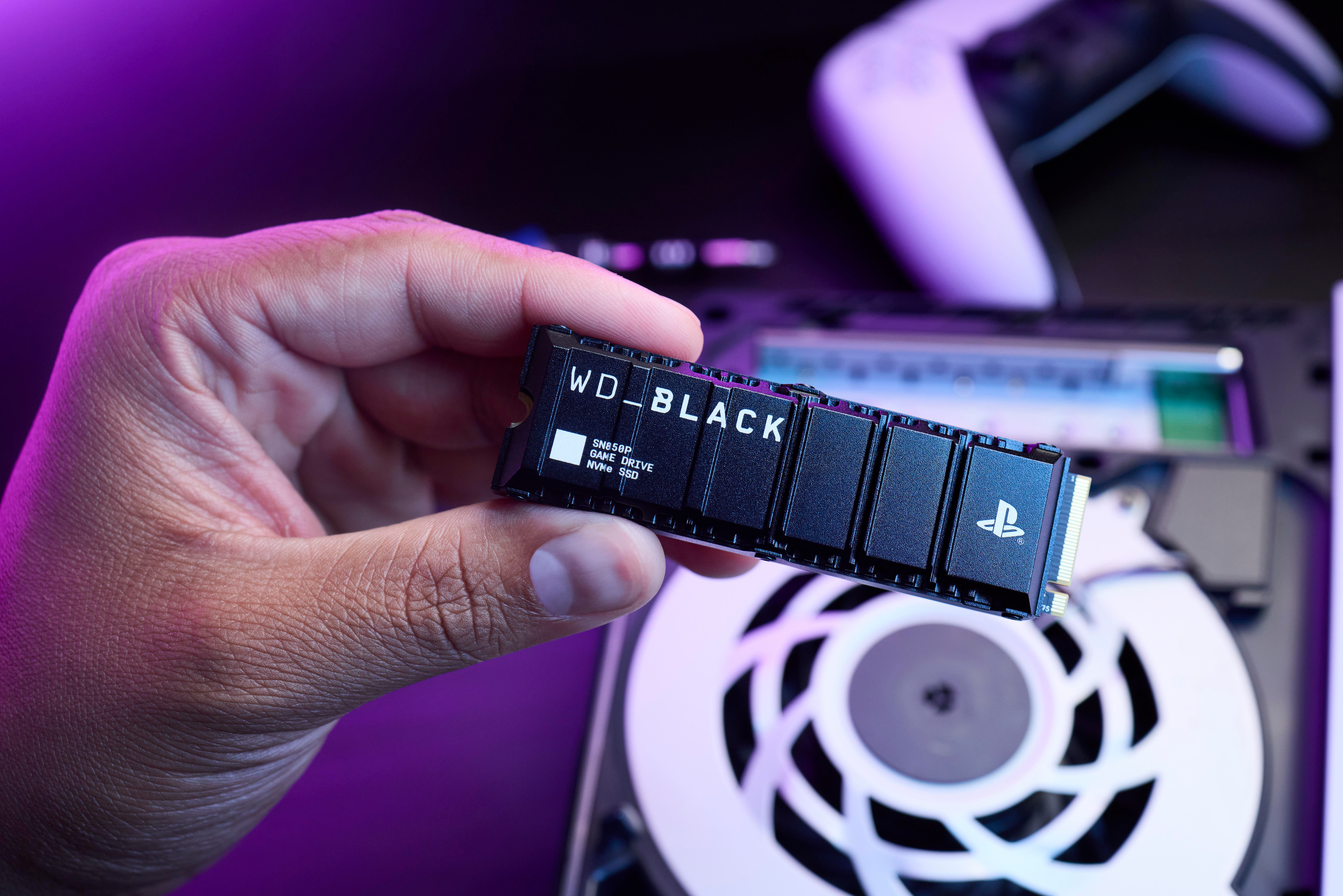 Hand hält eine schwarze WD_BLACK SSD, Spielkonsole und Controller im Hintergrund sichtbar.