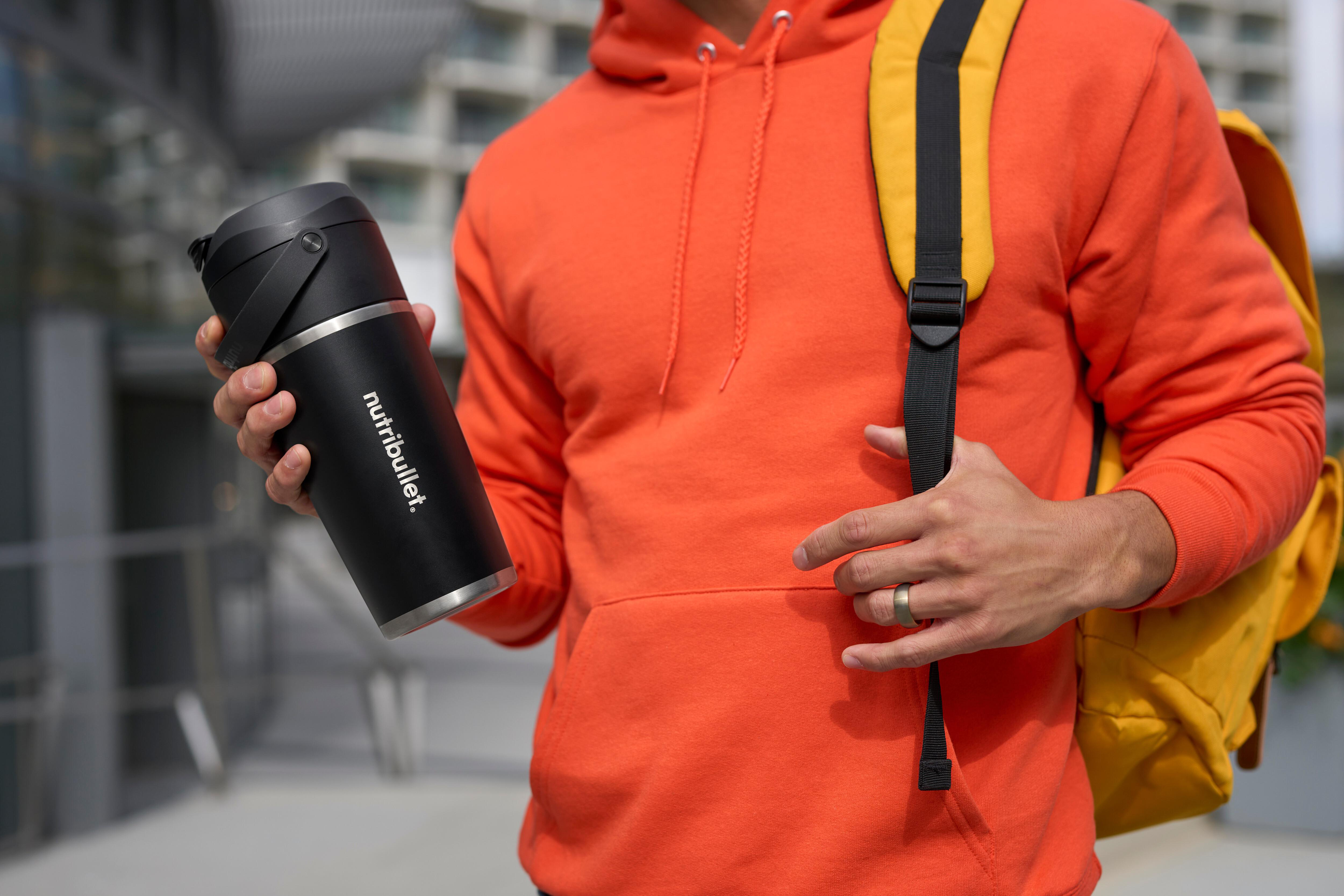 Person in orangefarbenem Hoodie, die eine schwarze Wasserflasche hält, gelber Rucksack im Hintergrund, draußen.
