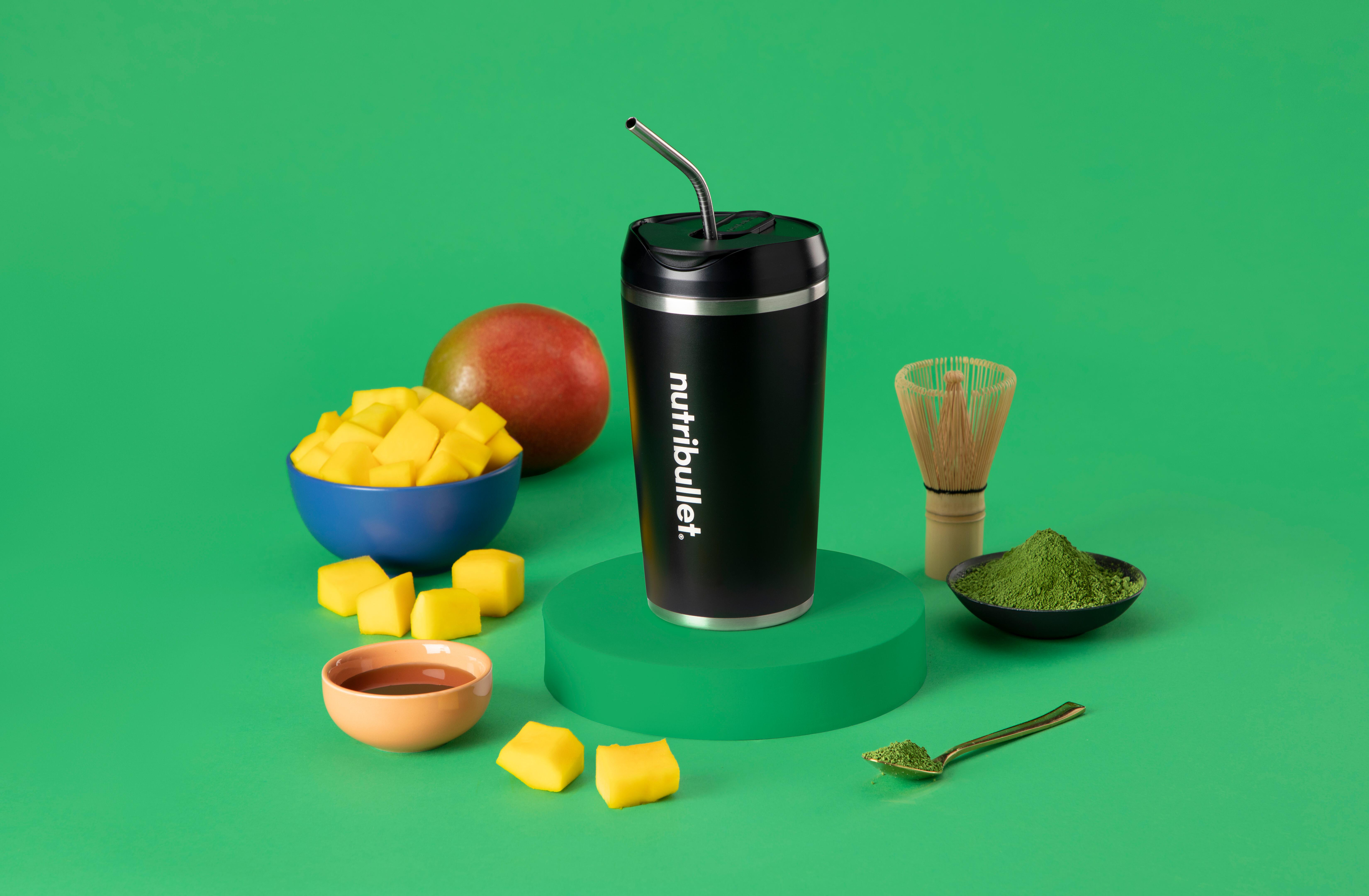 Ein schwarzer Becher mit Strohhalm, umgeben von Mangoscheiben, Matcha und einer Mango. Grüner Hintergrund.