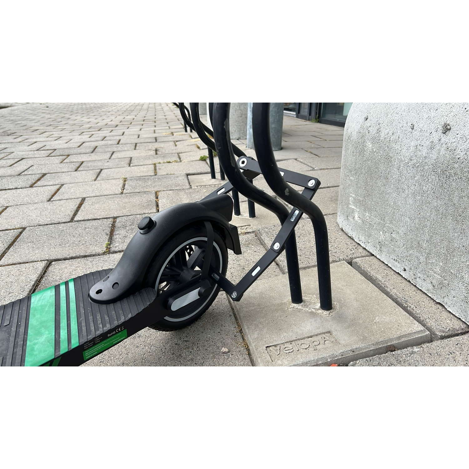 Trottinette électrique attachée à un porte-vélos en métal. Trottinette noire, plateau vert, pavé gris.