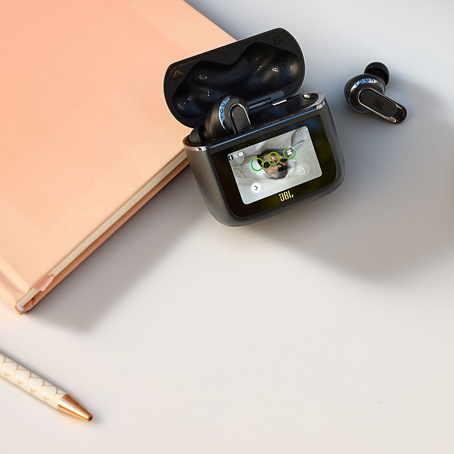 Earbuds in a charging case with a screen next to a notebook and a pen.