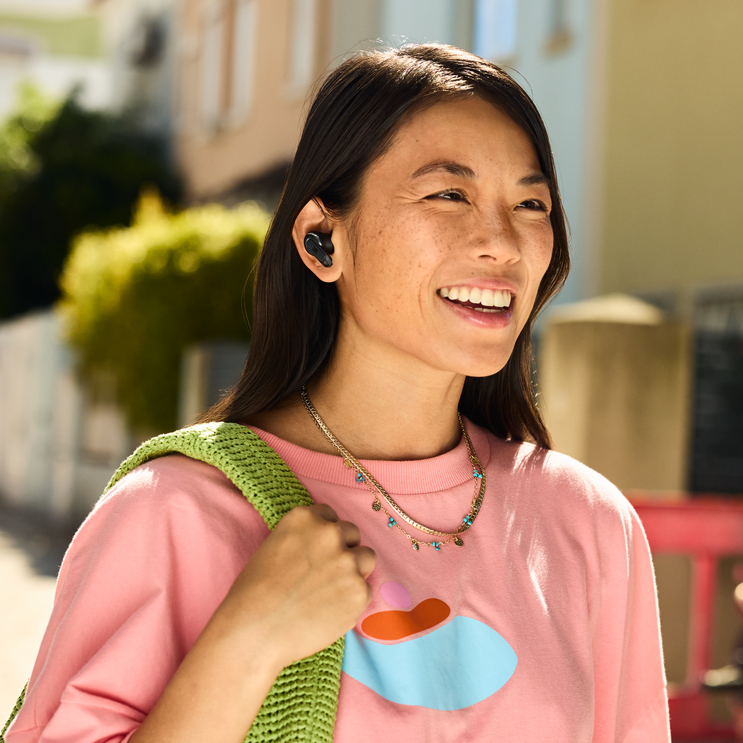 Smiling woman wearing black earbuds, walking on a street. Pink shirt, green bag, sunny day.