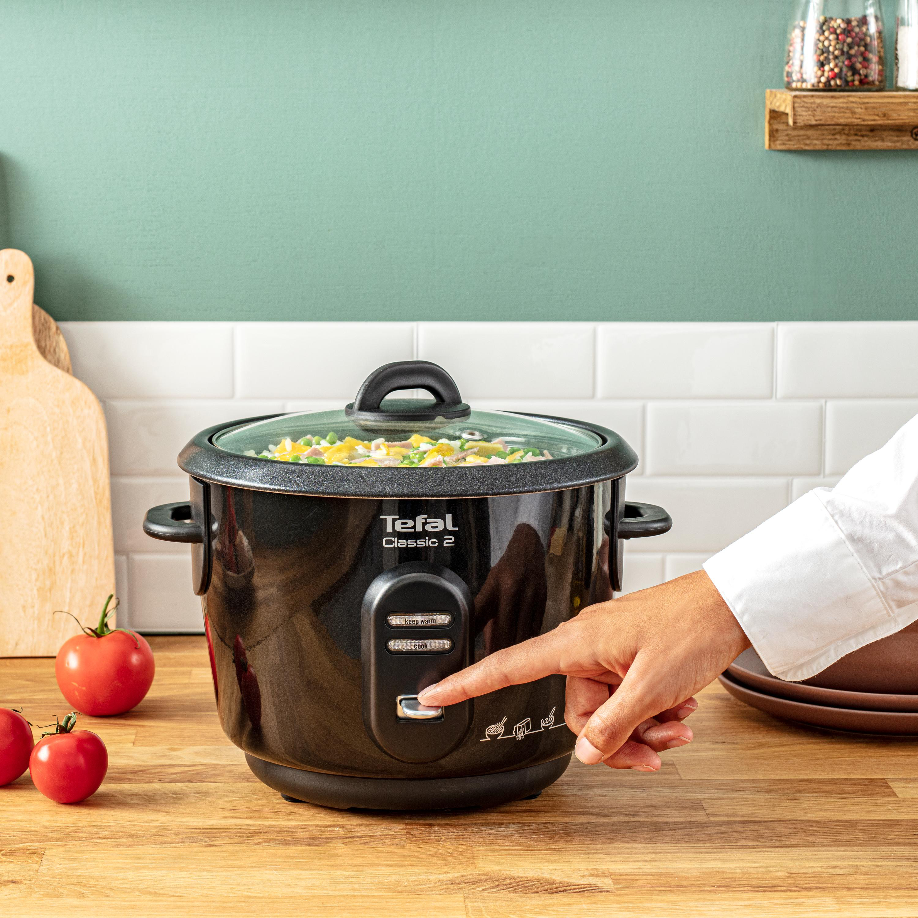 Schwarzer Reiskocher, Hand drückt Knopf. Tomaten und Schneidebrett im Hintergrund.