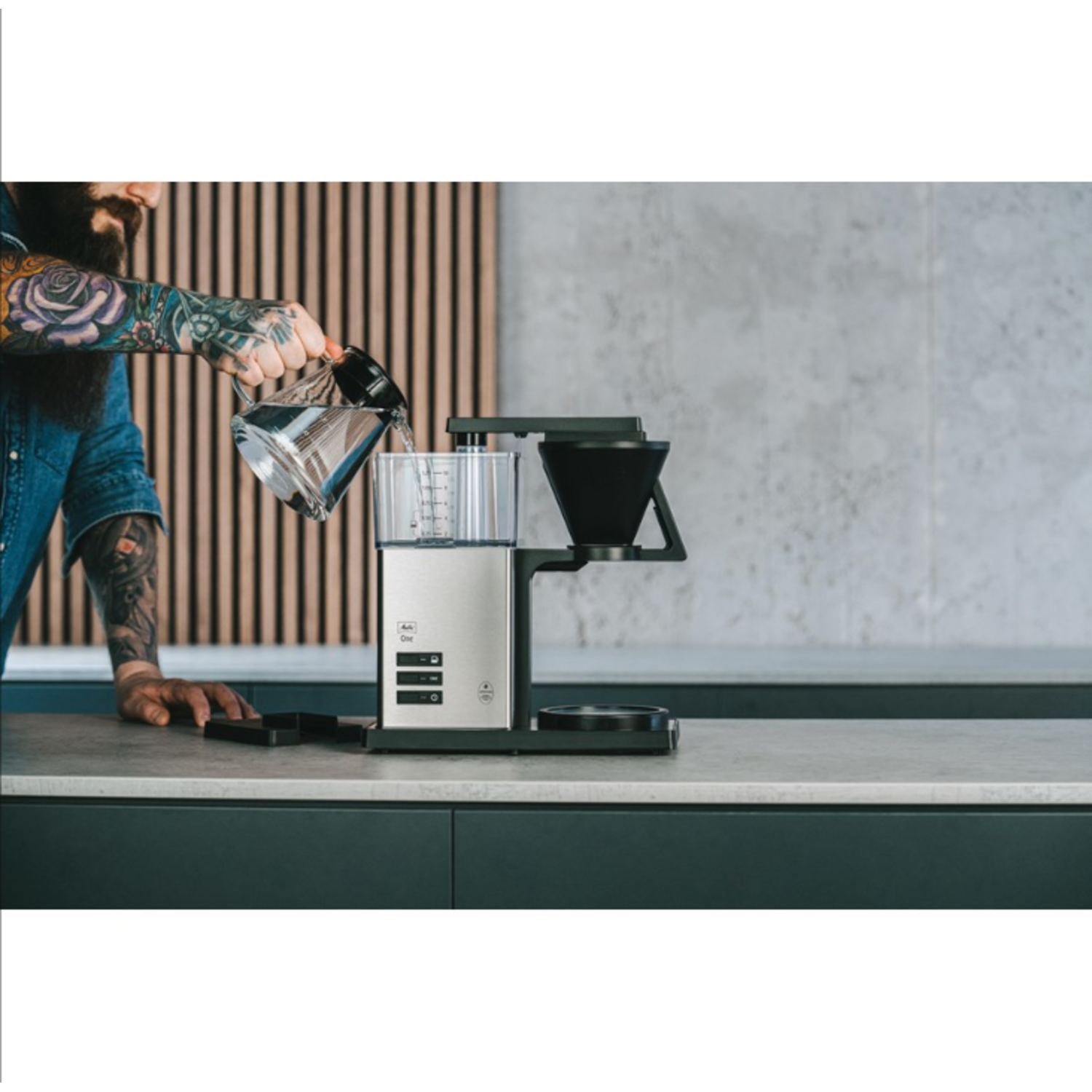 Een getatoeëerde man giet water in een koffiezetapparaat. Achtergrond: aanrecht en houten muur.