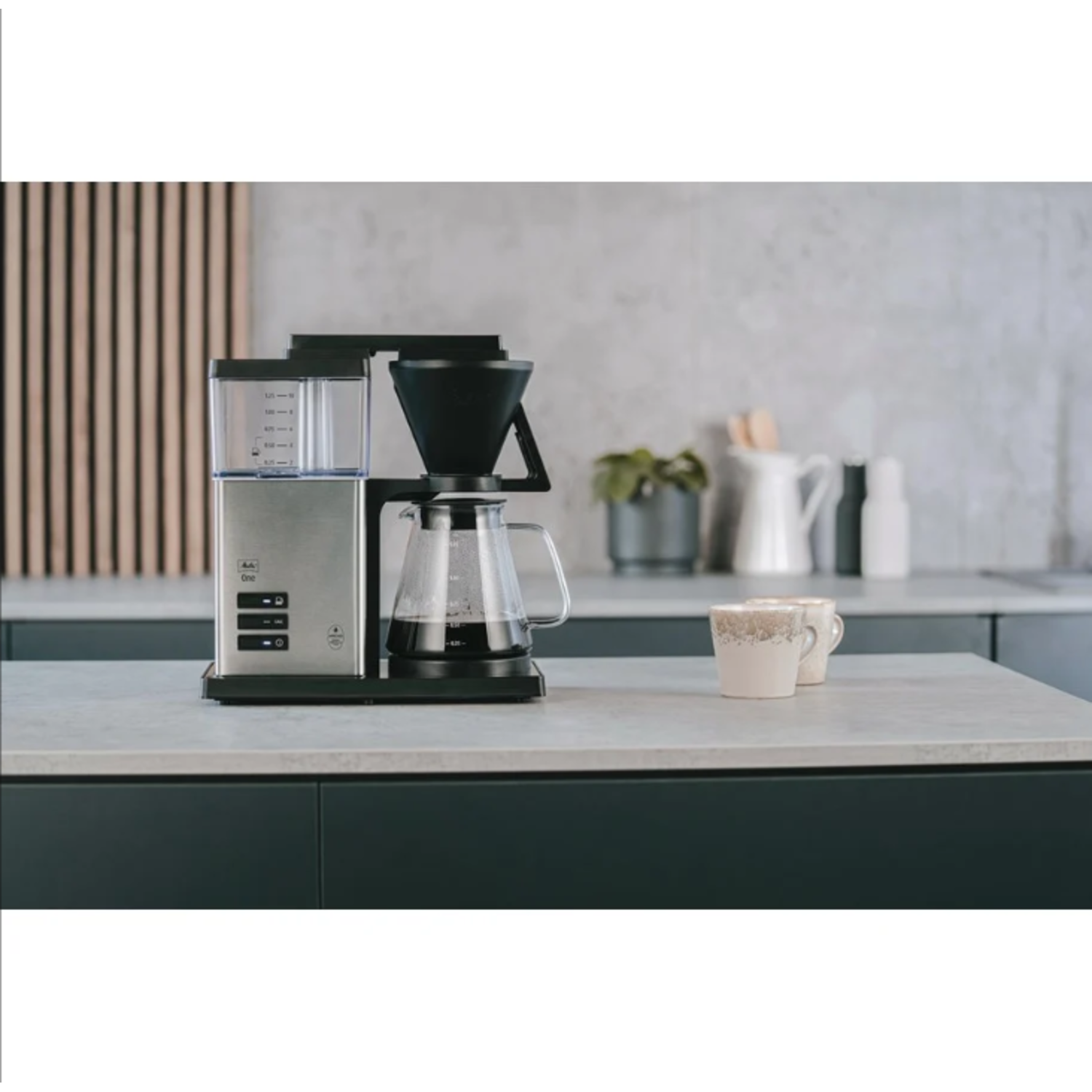 Een roestvrijstalen koffiezetapparaat met een glazen kan, op een aanrecht met twee kopjes.