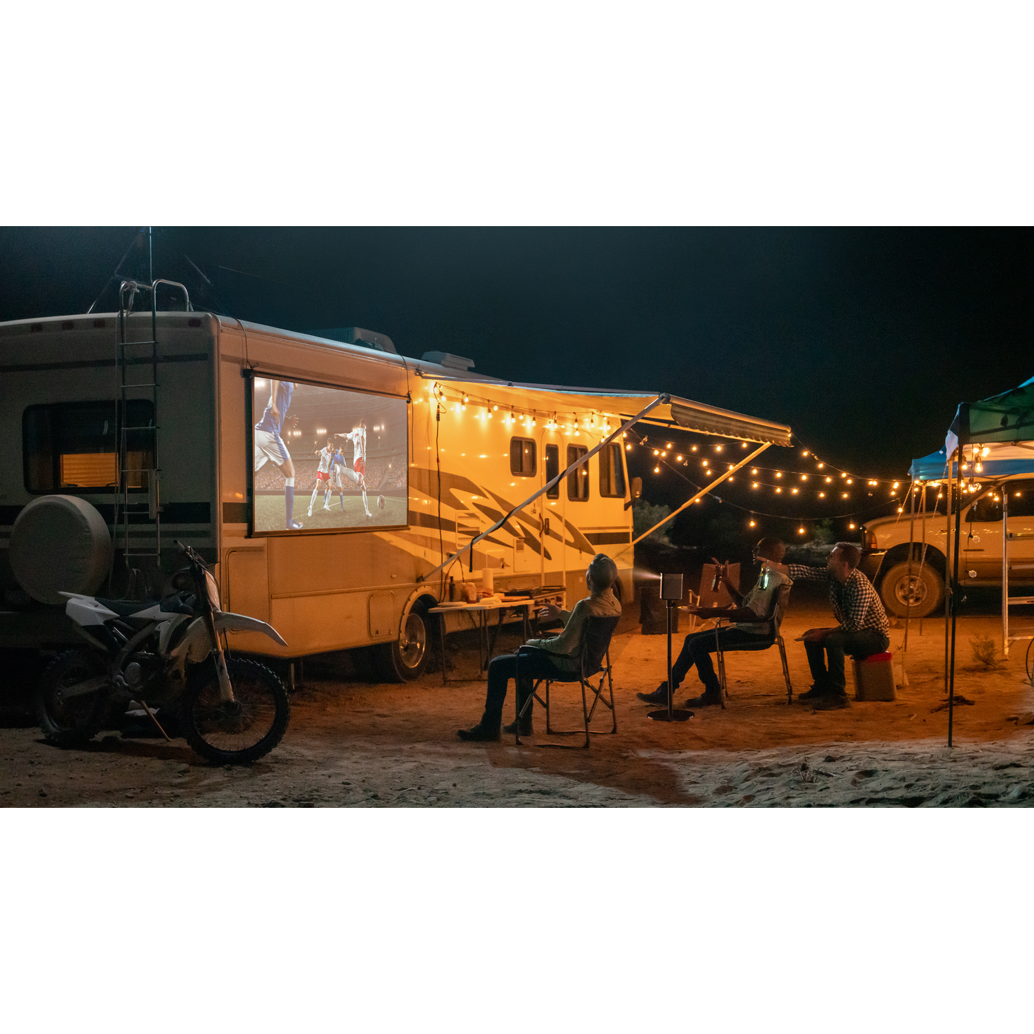 Groupe regardant un film sur écran sur un camping-car. Moto garée à proximité. Guirlandes lumineuses aussi.