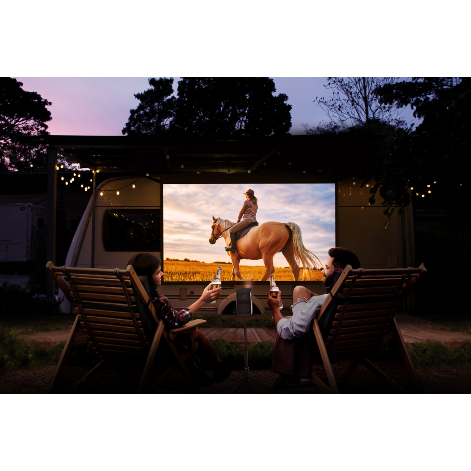 Couple regarde un film sur écran extérieur. Arbres, camping-car, ciel crépusculaire.