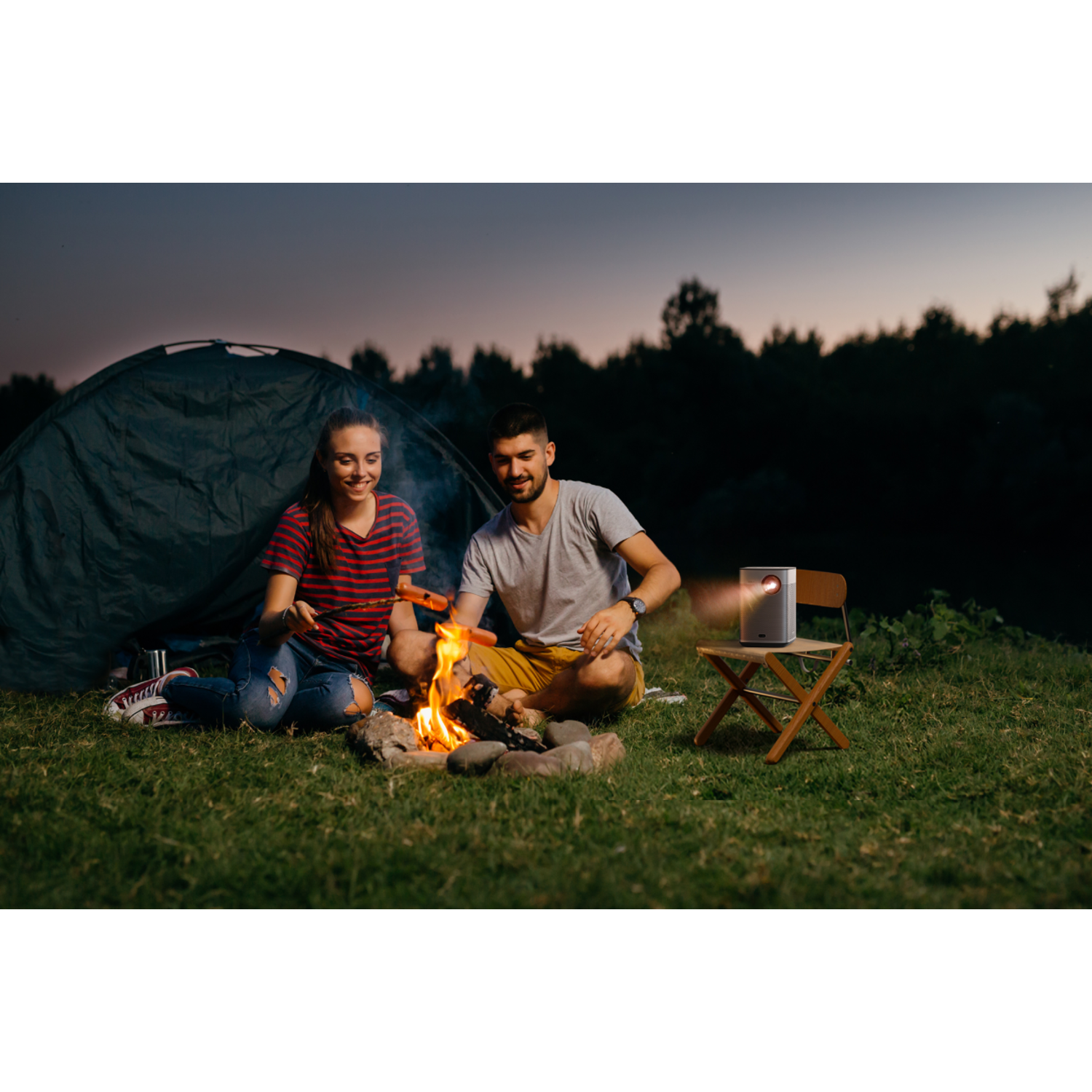 Un couple est assis près d'un feu de camp. Un projecteur affiche une image sur un support à proximité.