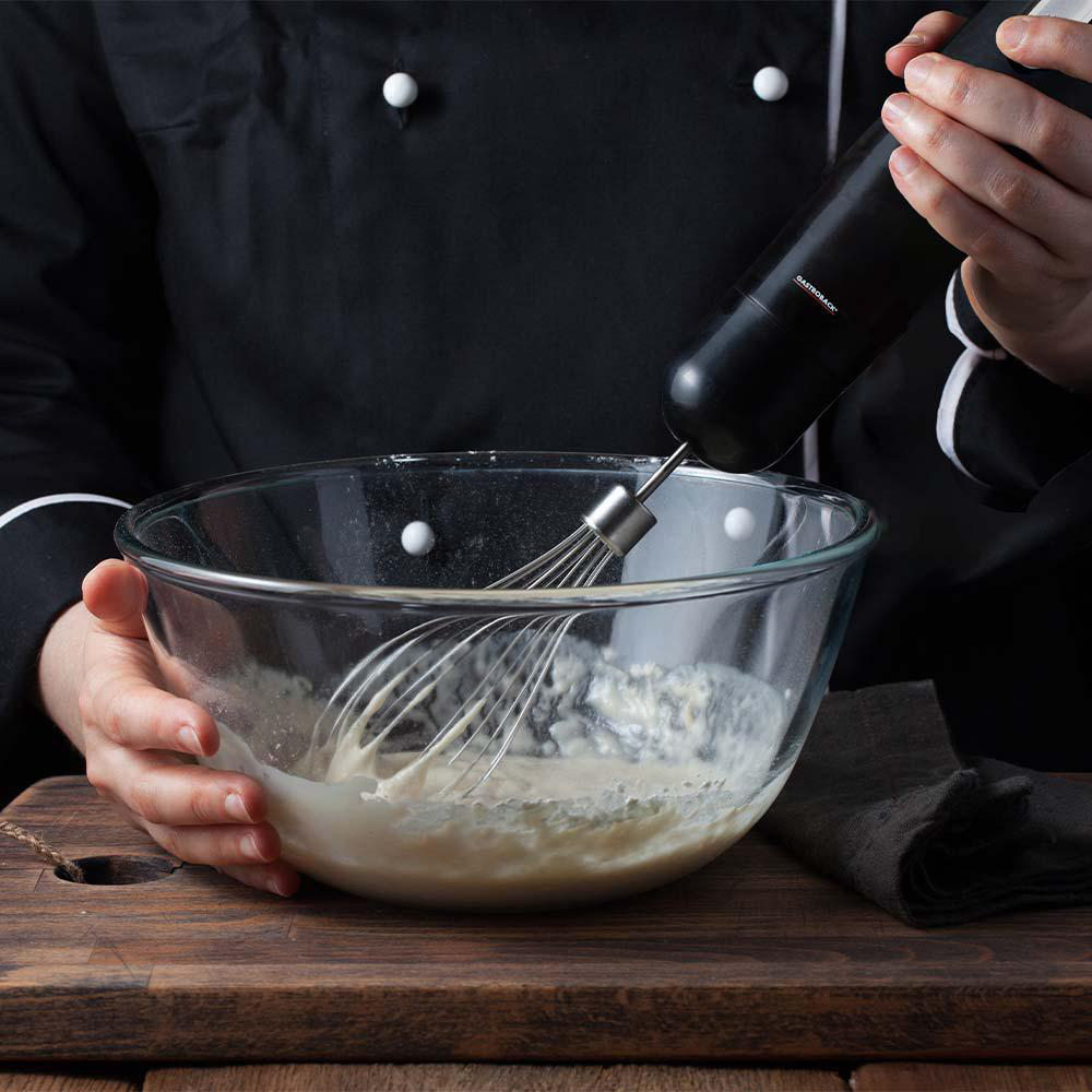 Ein Koch verquirlt Sahne in einer Glasschüssel mit einem Handmixer auf einem Holzbrett.