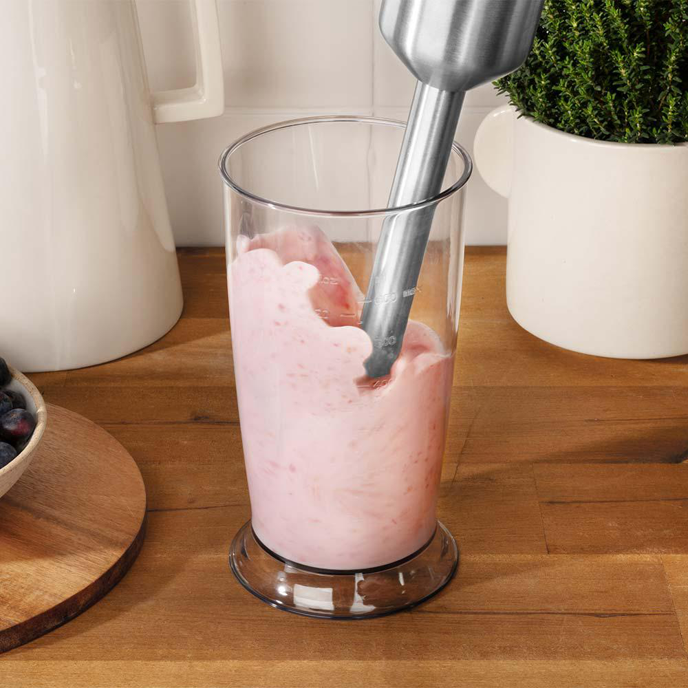 Ein Stabmixer ist in einem Glas mit rosa Flüssigkeit, mit weißem Krug und Pflanze im Hintergrund.
