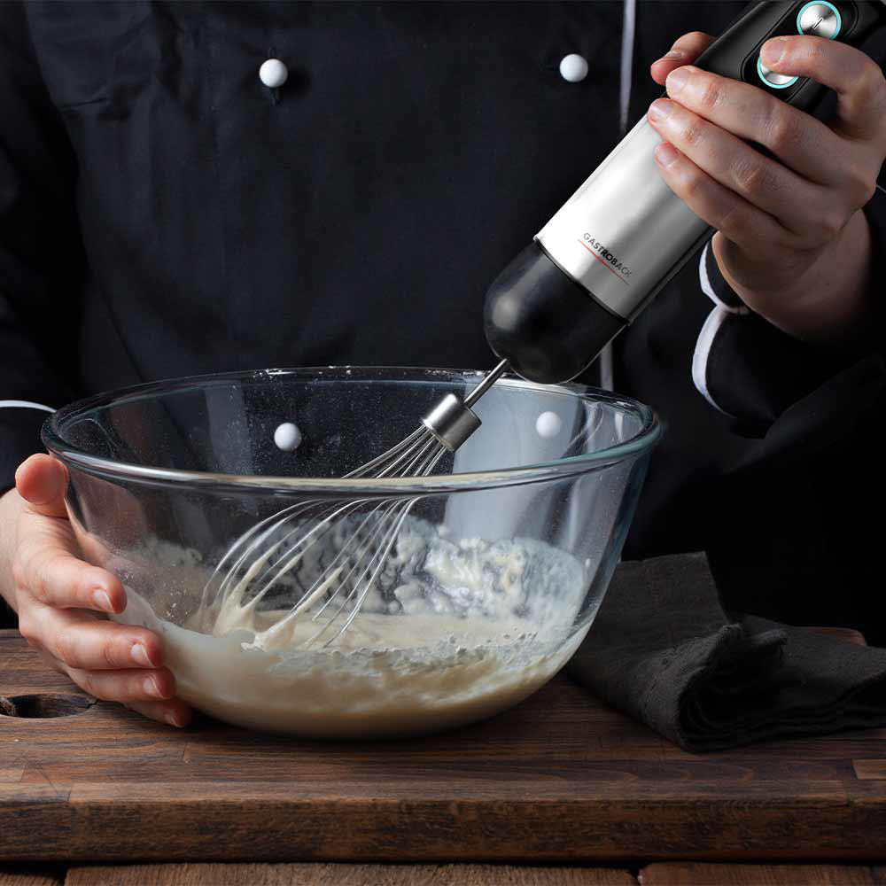 Person benutzt einen silbernen und schwarzen Handmixer in einer Glasschüssel. Holzoberfläche.